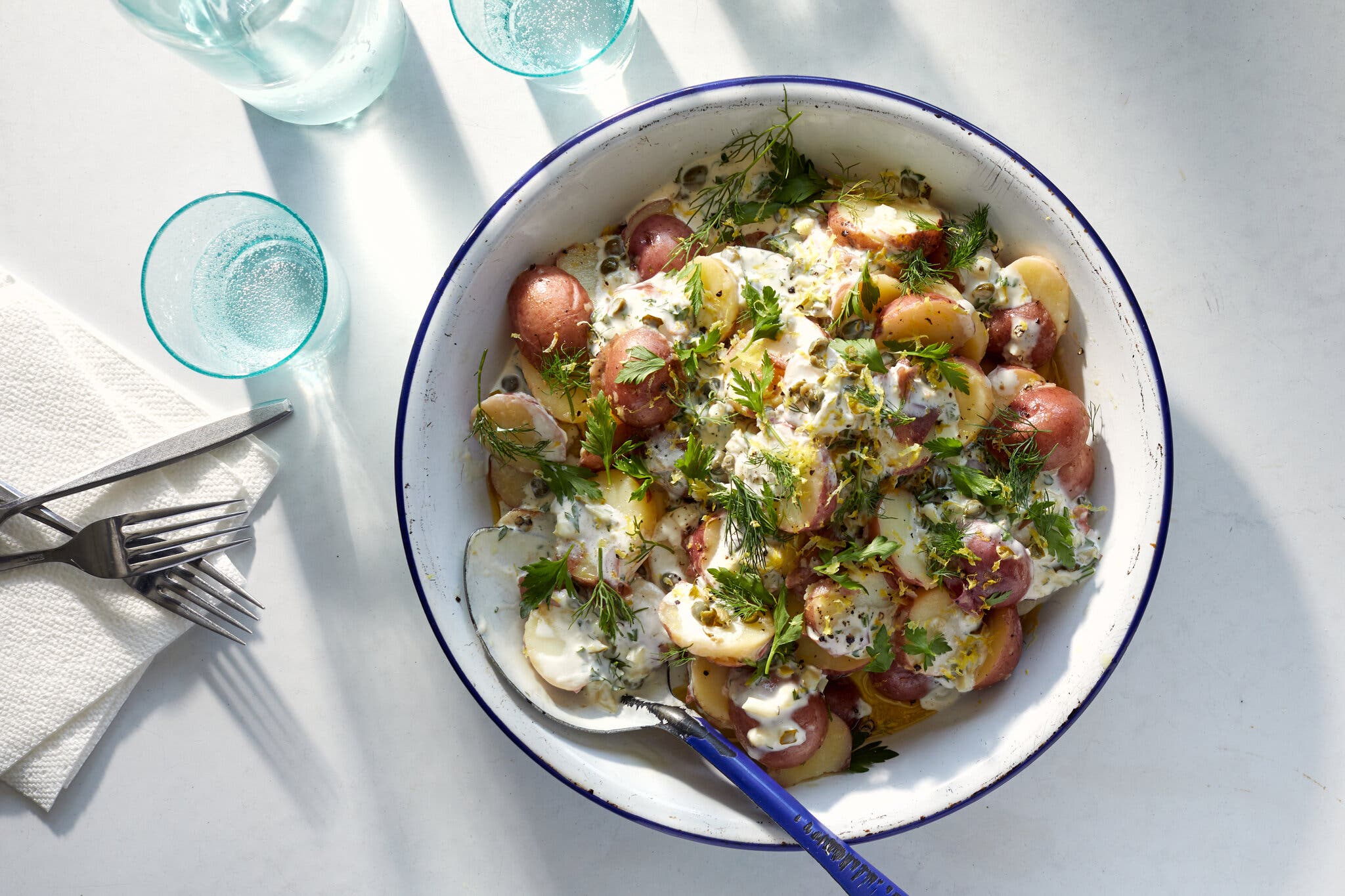 A white enamel serving bowl with a blue edge holds a salad of tiny red potatoes, creamy dressing and herbs. Light blue glasses hold sparkling water.