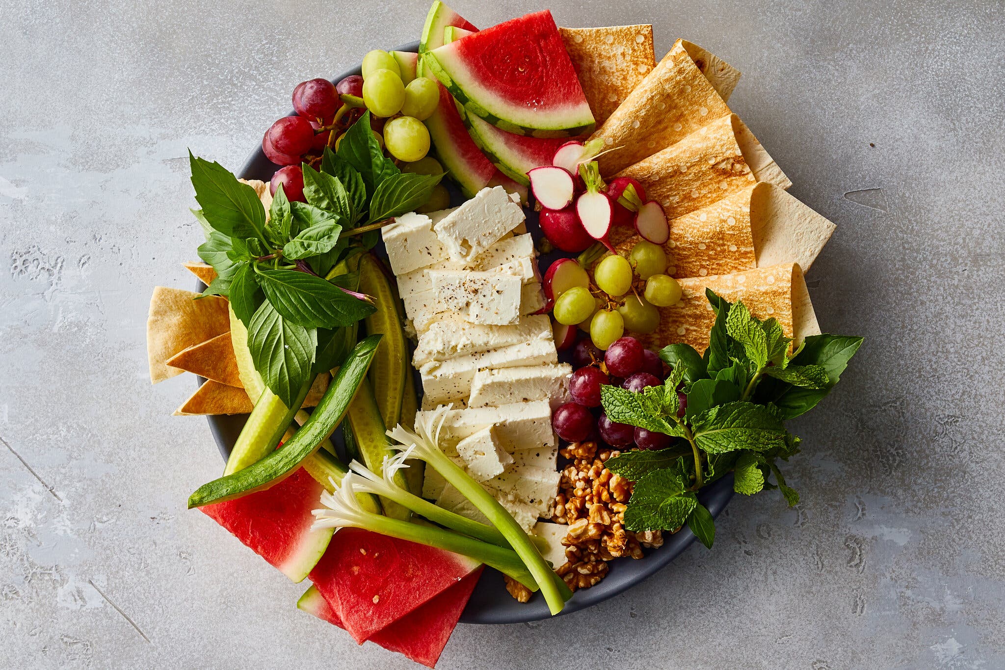 A platter teeming with watermelon, scallions, cucumbers, herbs, grapes, lavash, walnuts and feta.