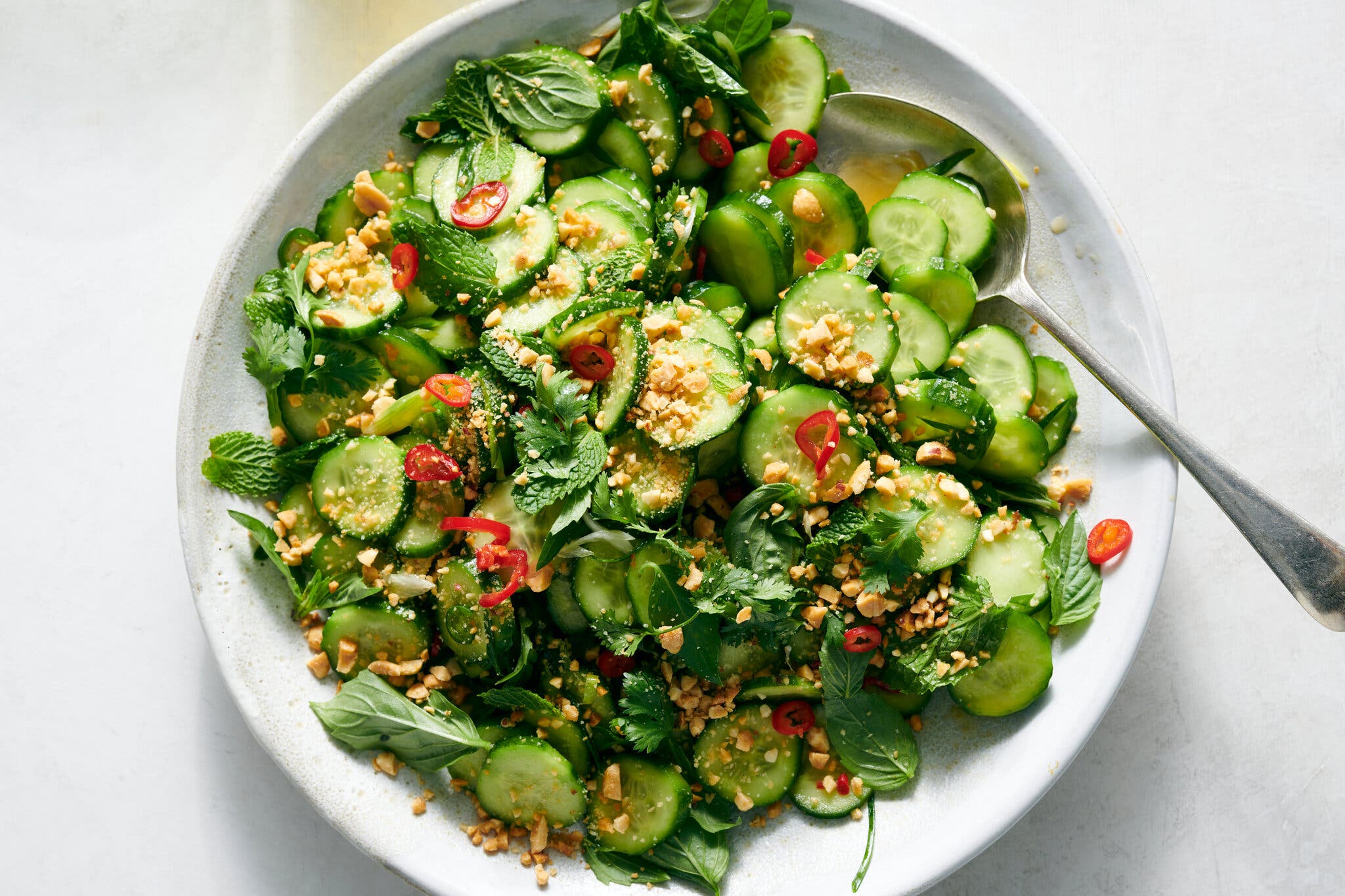 A salad of cucumbers, chiles, peanuts and mint rests in a shallow white bowl with a spoon tucked in.