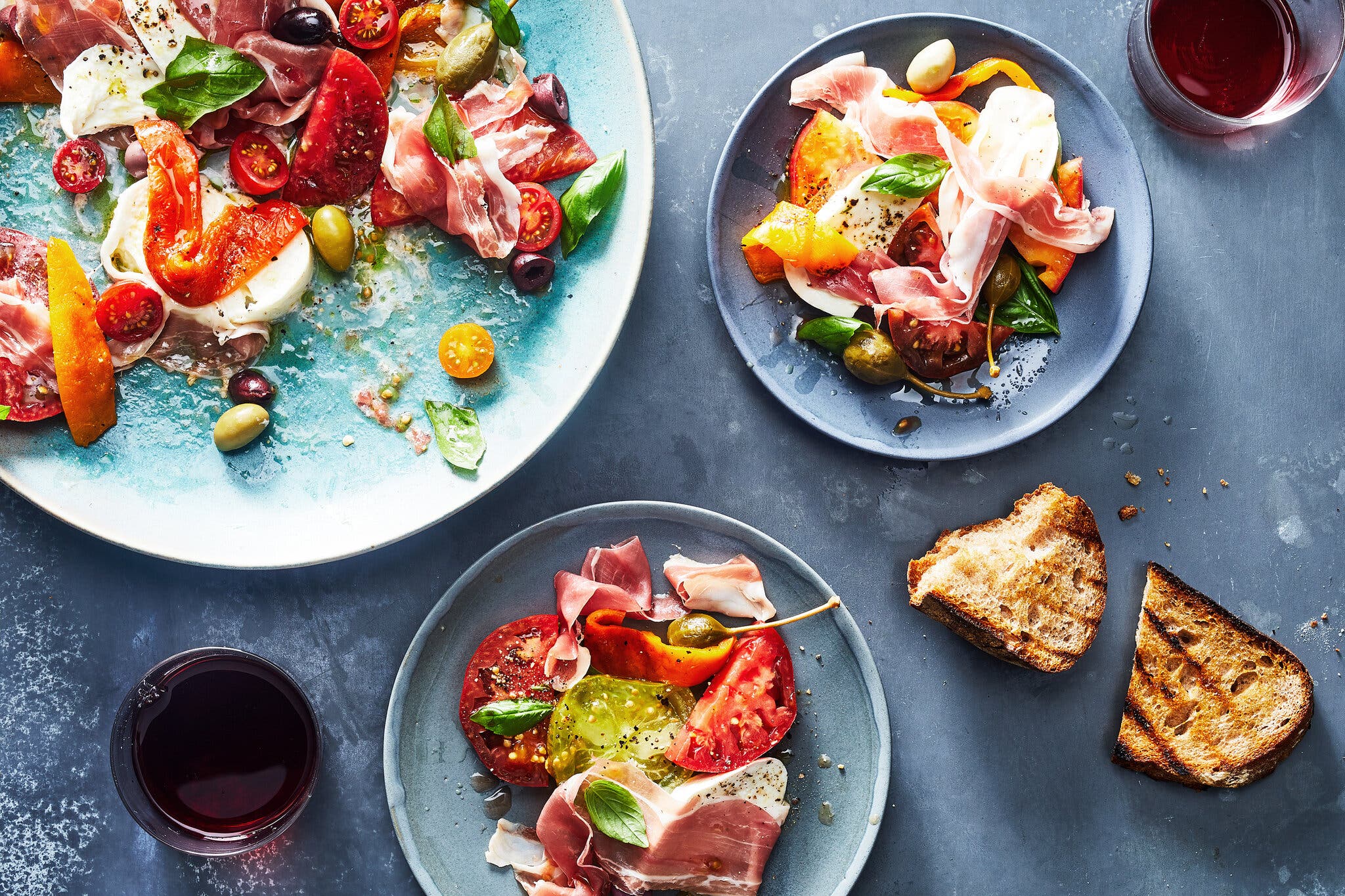 A platter holding tomatoes, mozzarella, prosciutto and olives peeks into the frame next to two servings.