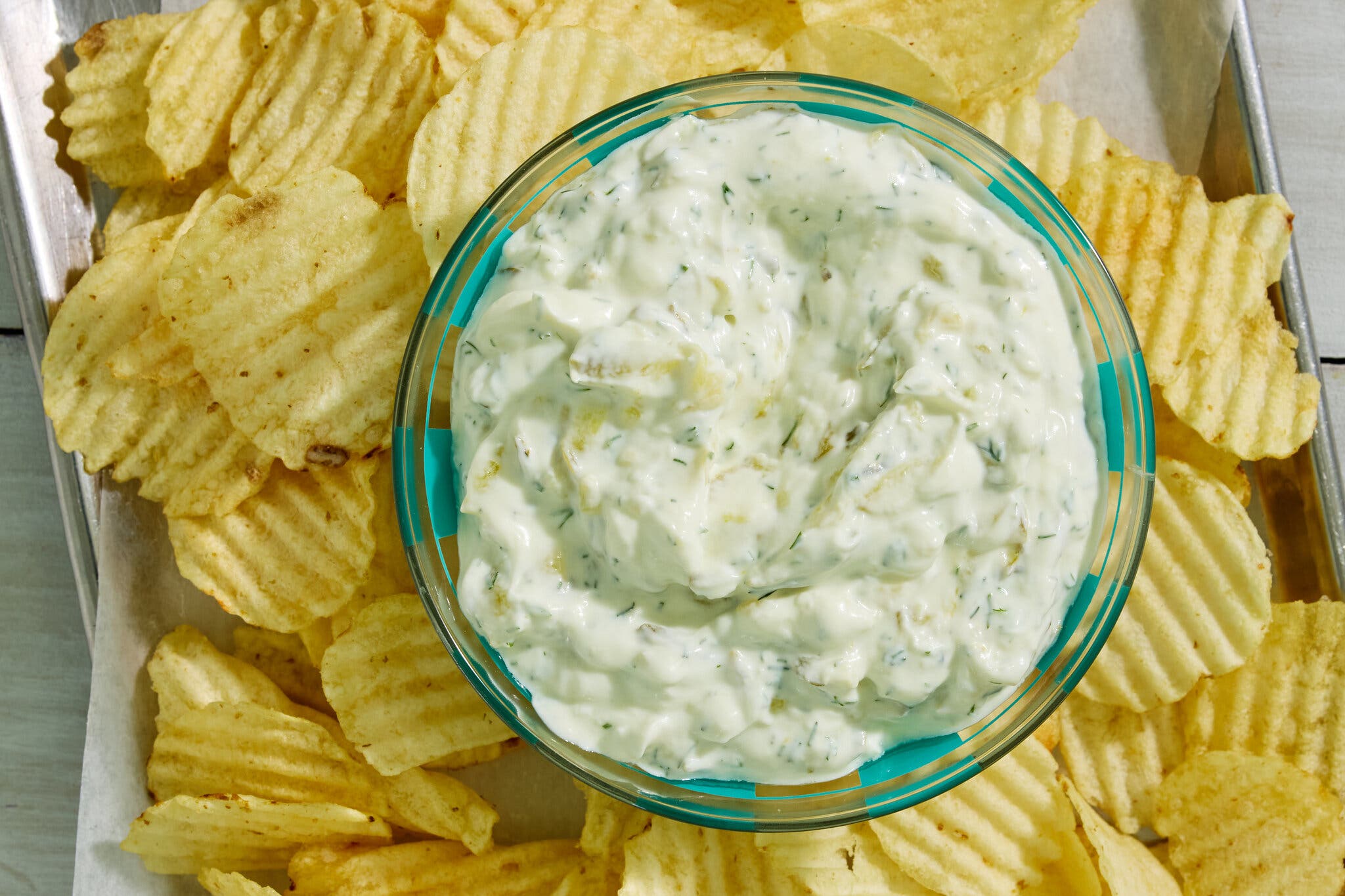A glass dish of creamy pickle dip rests on a tray full of ridged potato chips.