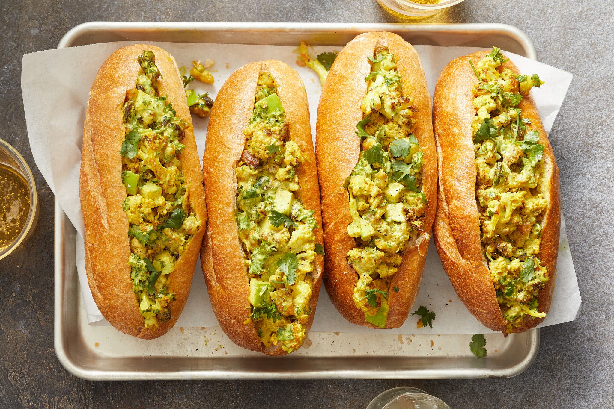 Four hoagie rolls split and stuffed with a marigold cauliflower salad rest on a parchment-lined sheet pan.