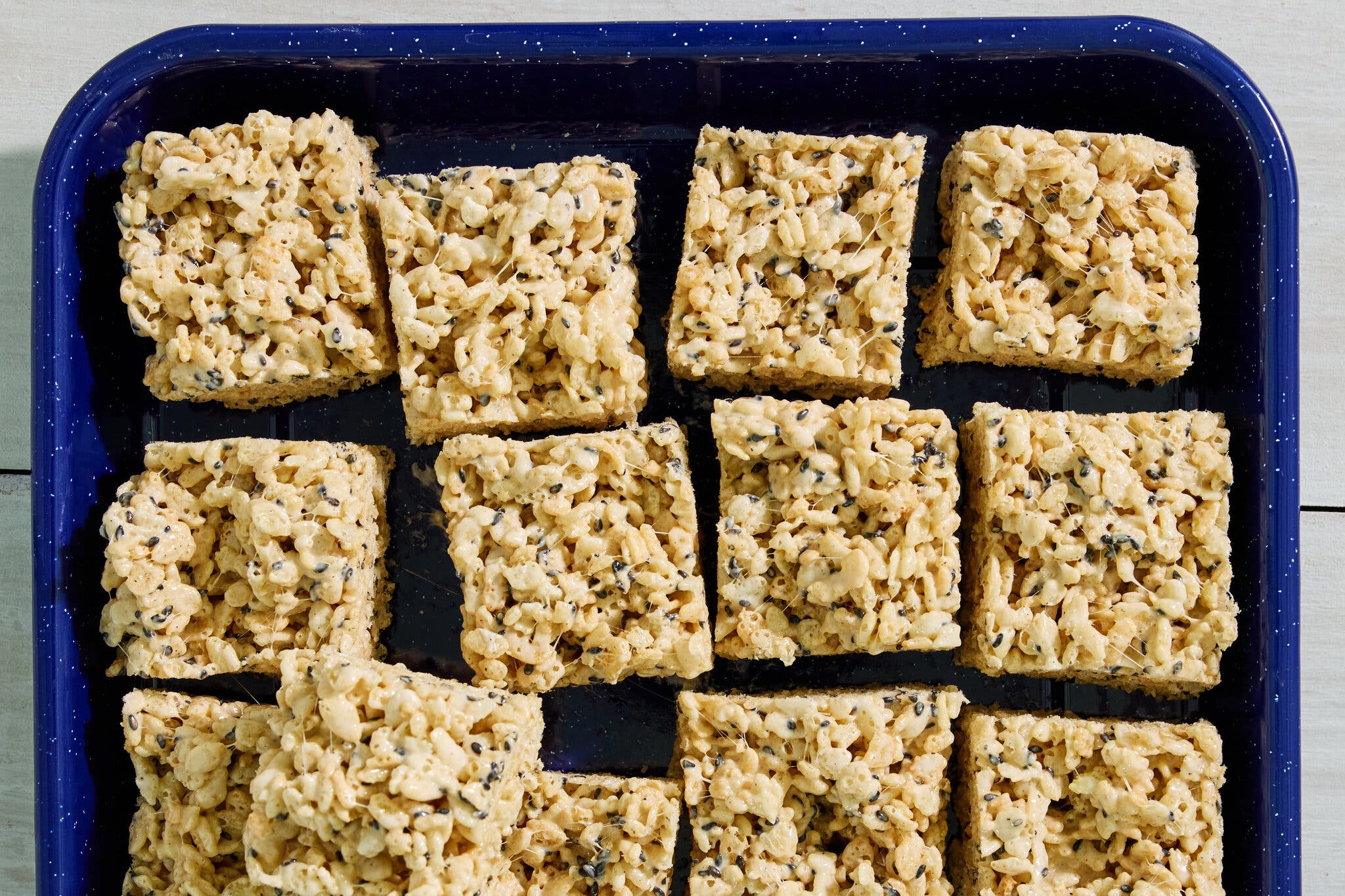 A dozen squares of Rice Krispies treats, speckled with black sesame, sit in a dark blue pan.