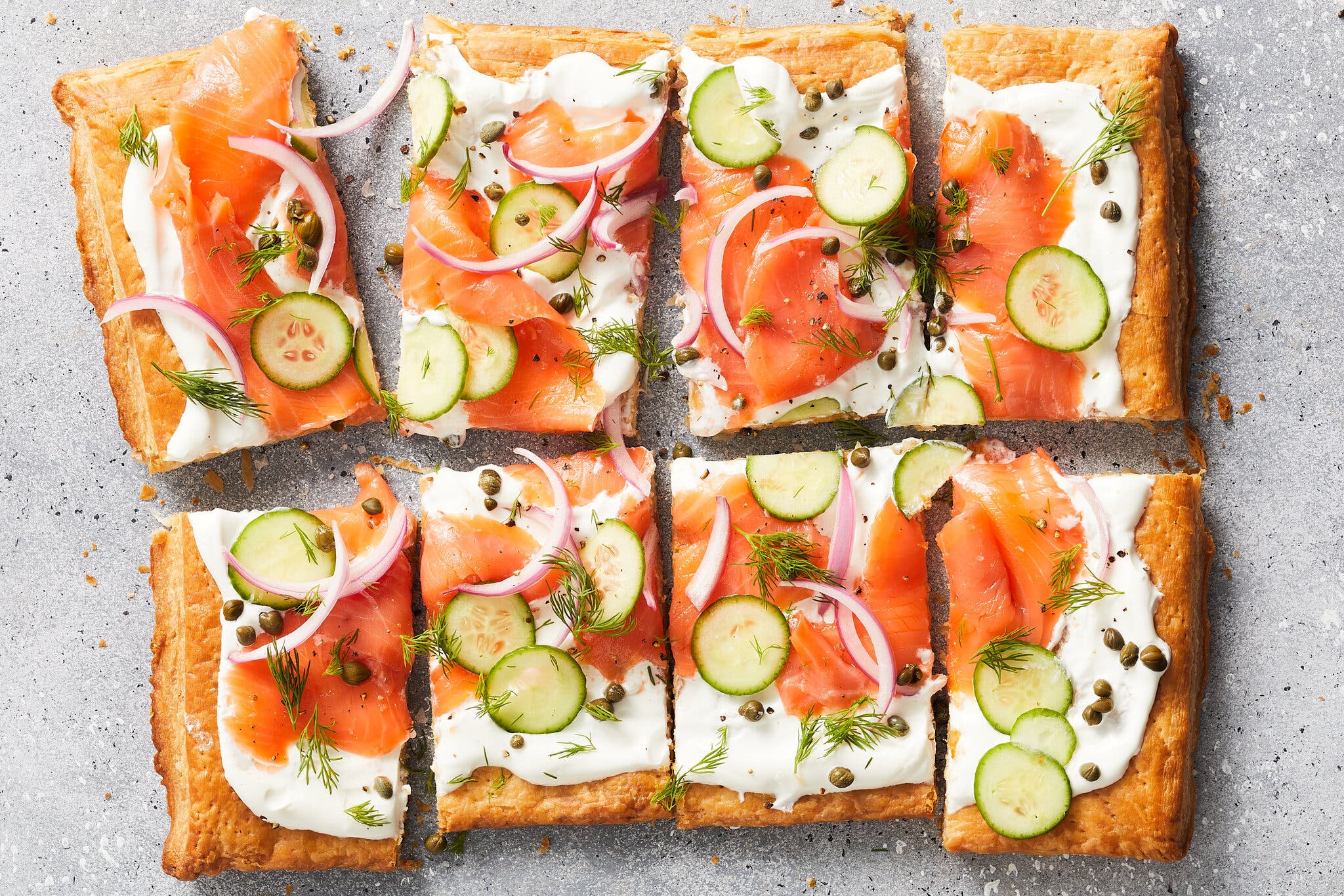 An overhead image shows a puff pastry tart that has been garnished with sour cream, smoked salmon, cucumbers, red onion, capers and dill.
