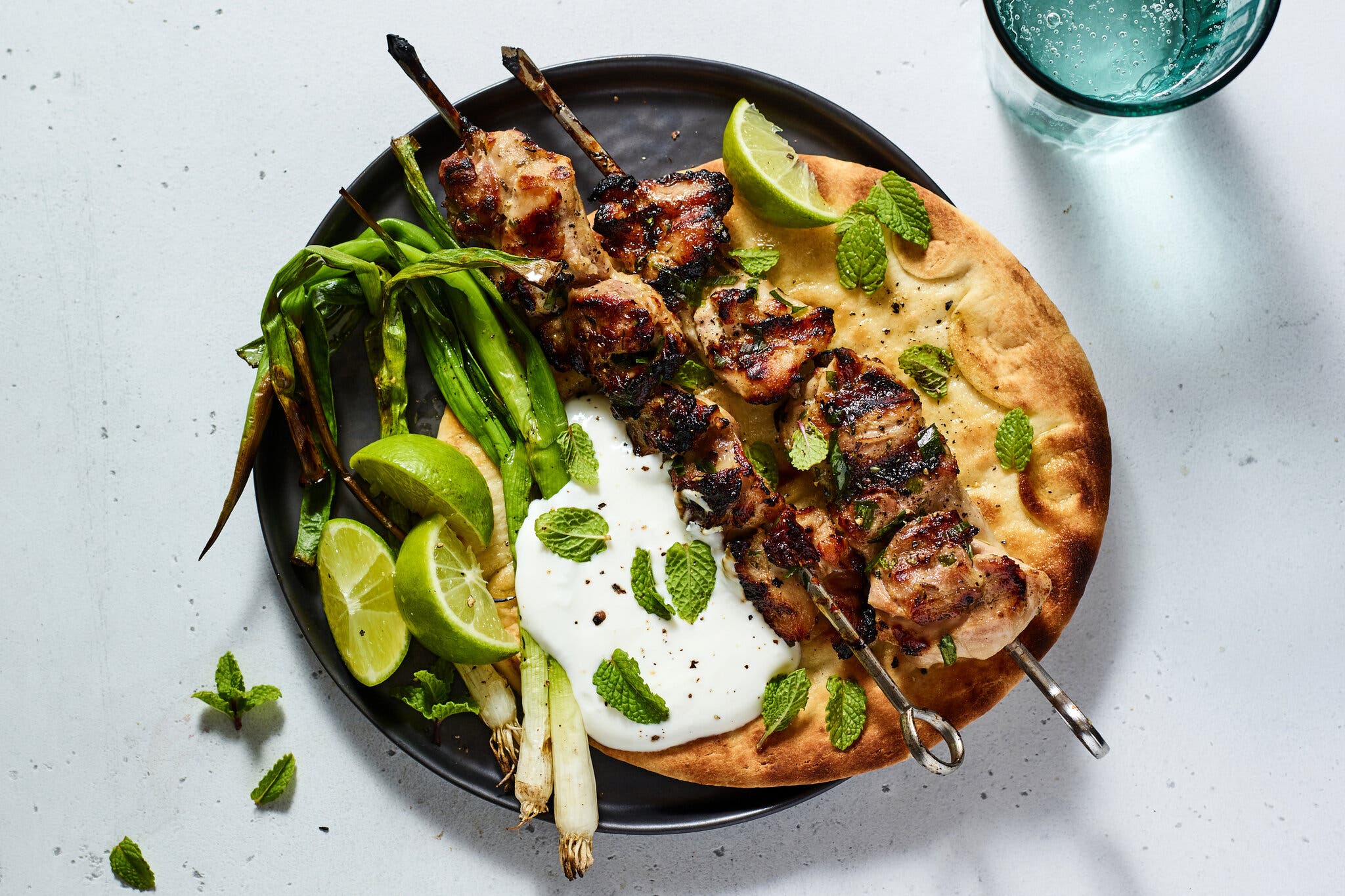 Two skewers of well-charred chicken pieces rest on a grilled flatbread, garnished with grilled scallions, yogurt sauce, mint and lime wedges.