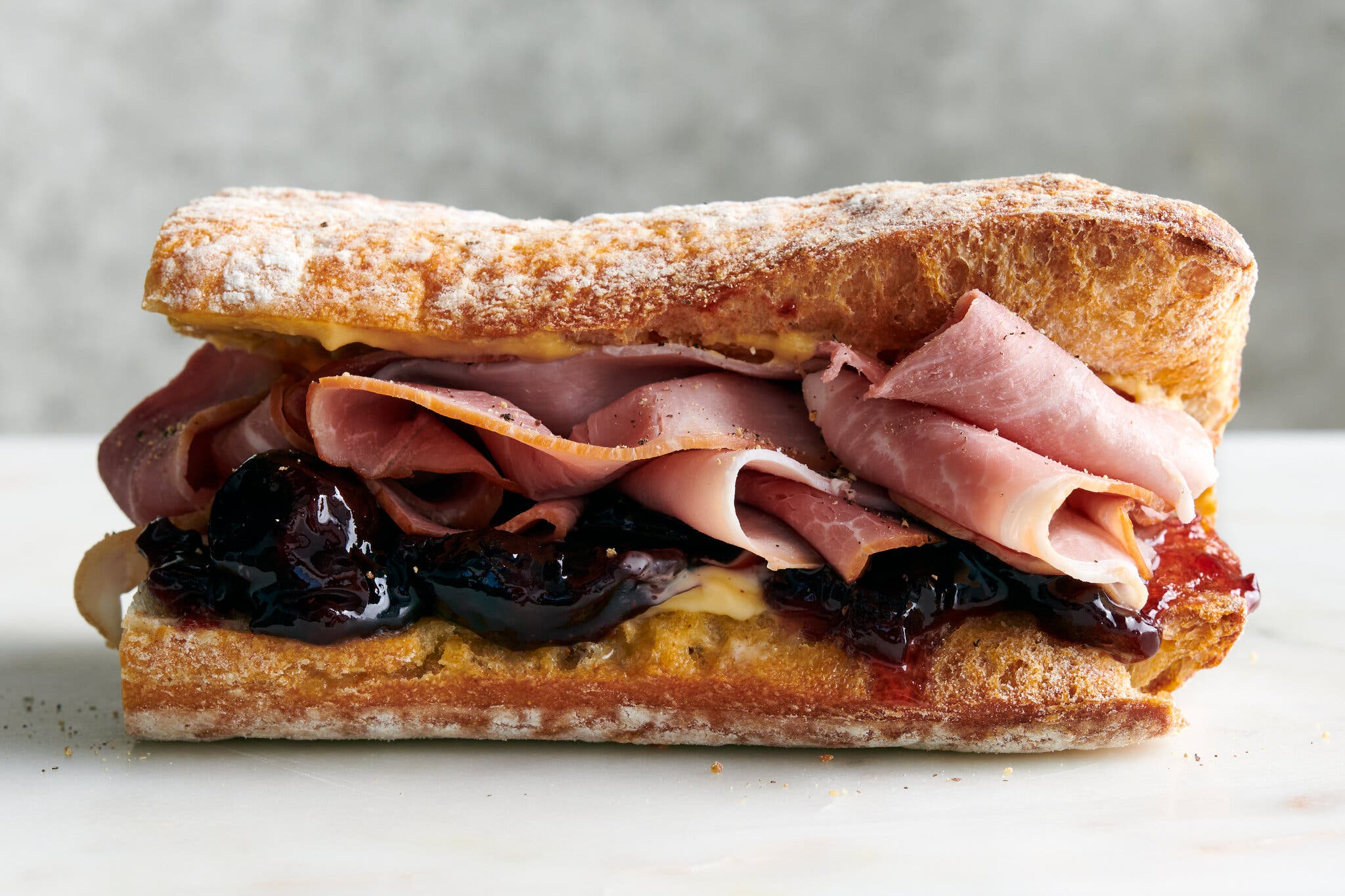 A side image of a baguette cut in half and filled with ham, rich cherry jam and butter.