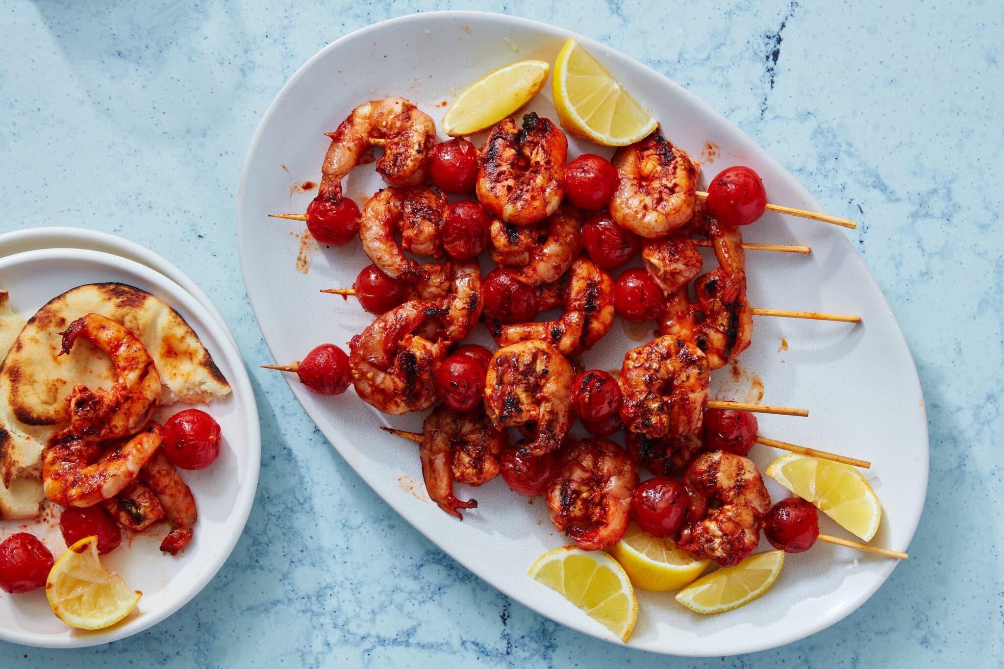 A white platter holds skewers of reddish grilled shrimp and cherry tomatoes. Lemon wedges are scattered around the plate.