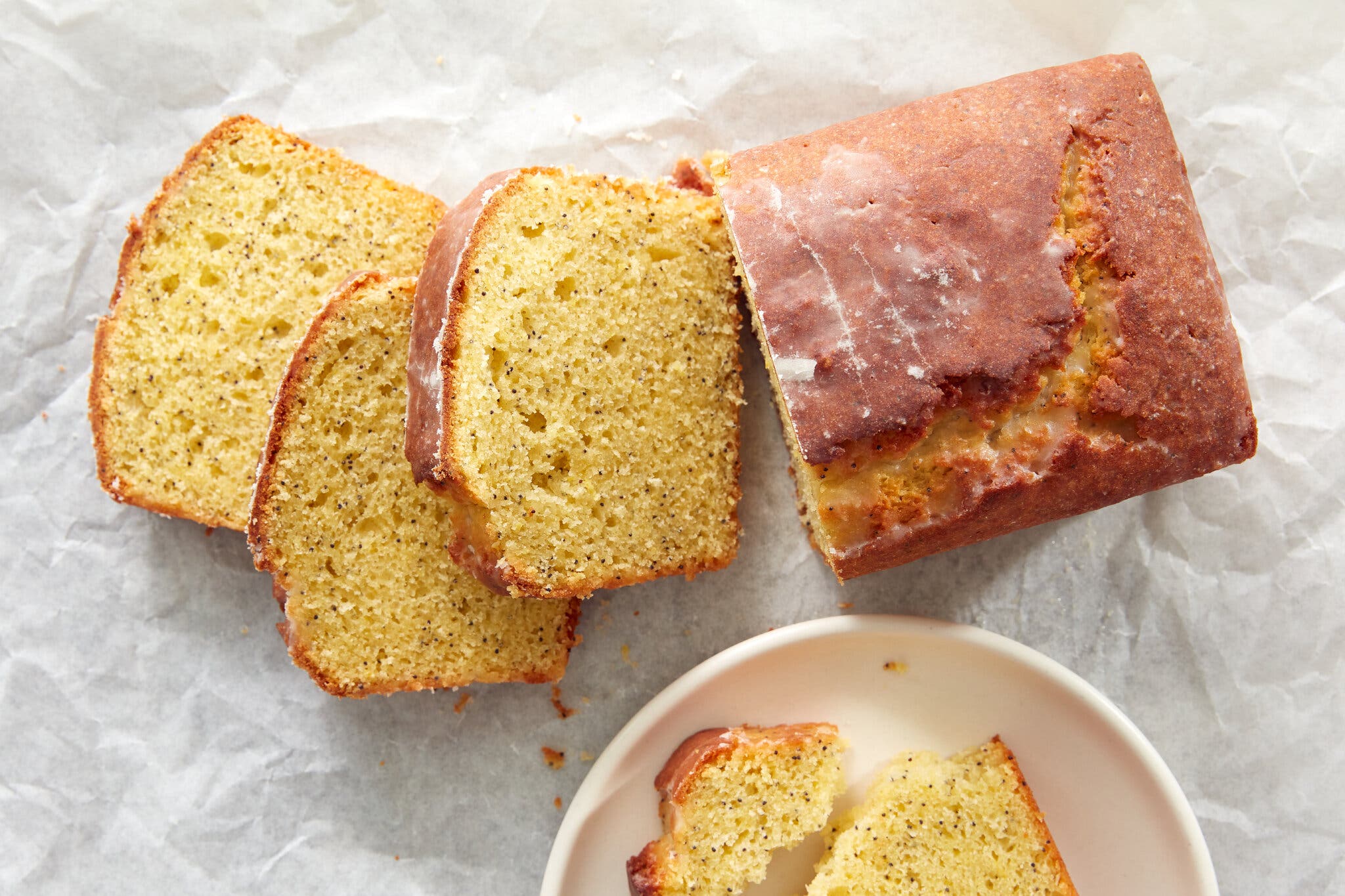A glazed partly sliced pound cake sits on a crumbled sheet of parchment. A single serving sits off to the side.