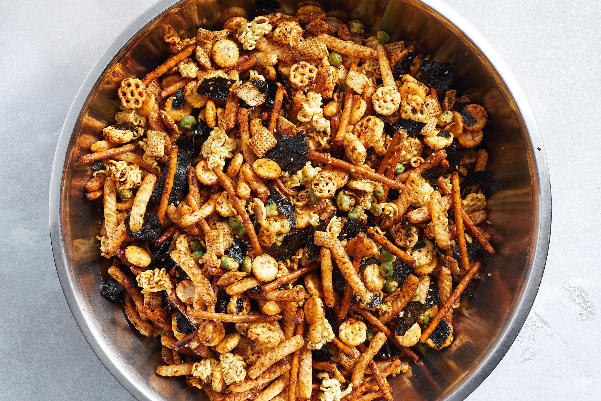 An overhead image of a snack mix with shrimp crackers, waffled rice cereal, pretzel sticks and toasted nori in a large steel bowl.