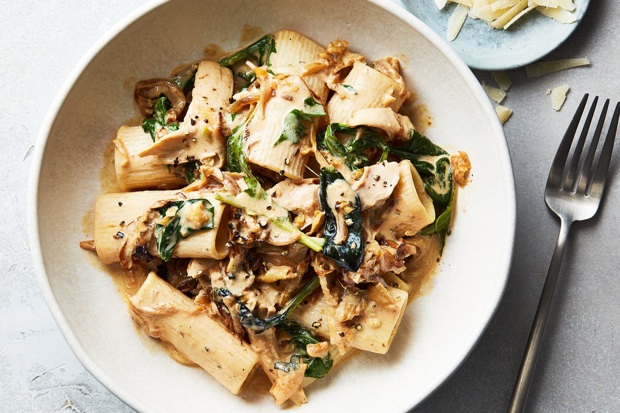 A creamy bowl of pasta topped with greens sits in a bowl. Shaved Parmesan is visible on the side.