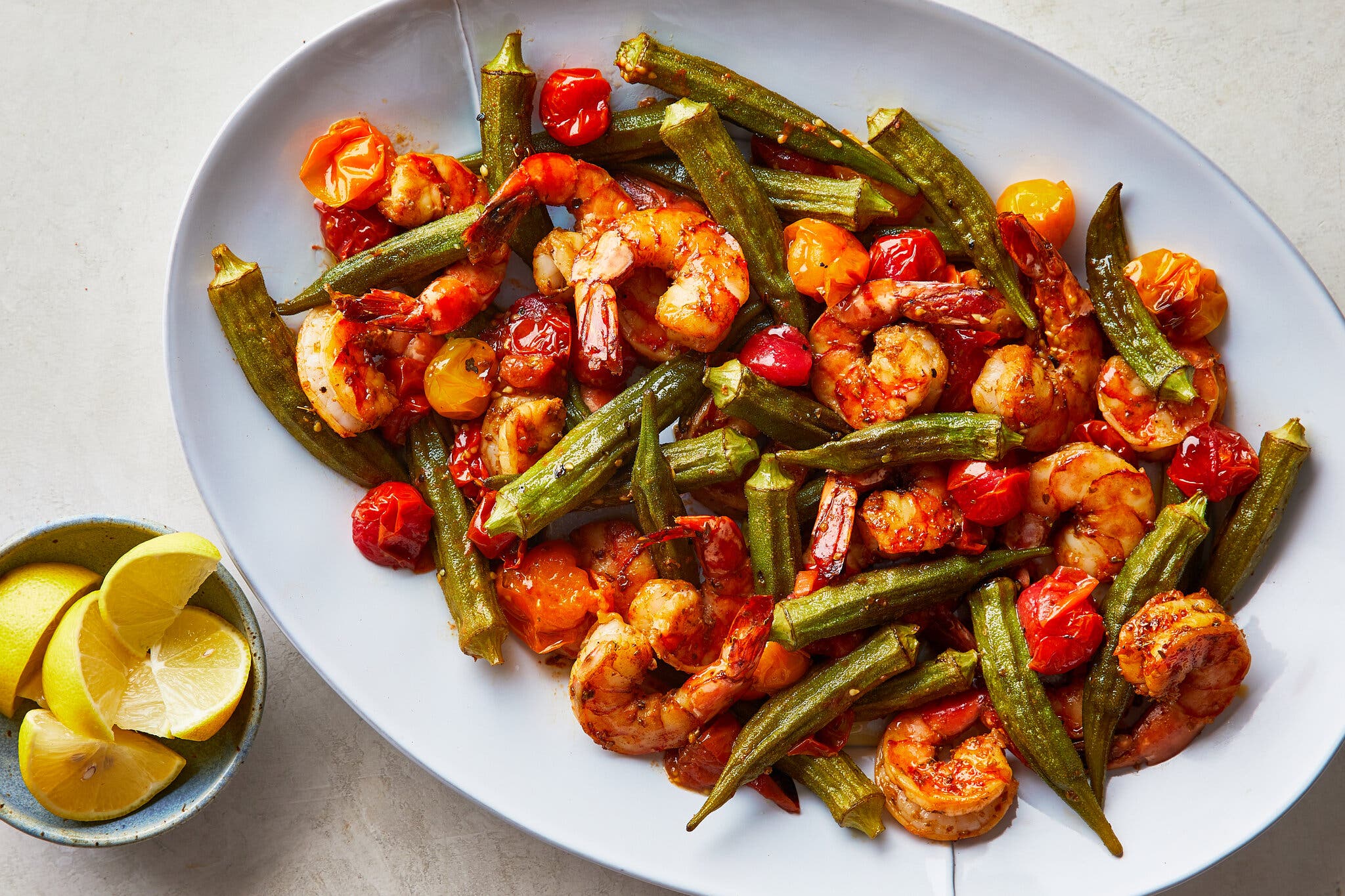 An overhead image of okra, tomatoes and shrimp on a white platter next to a small bowl of lemon wedges.