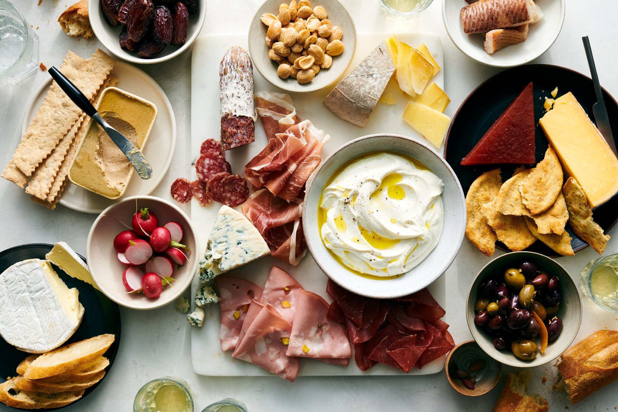 An overhead image of a charcuterie board spread with cheeses and meats. At the center is a bowl of whipped ricotta topped with olive oil.