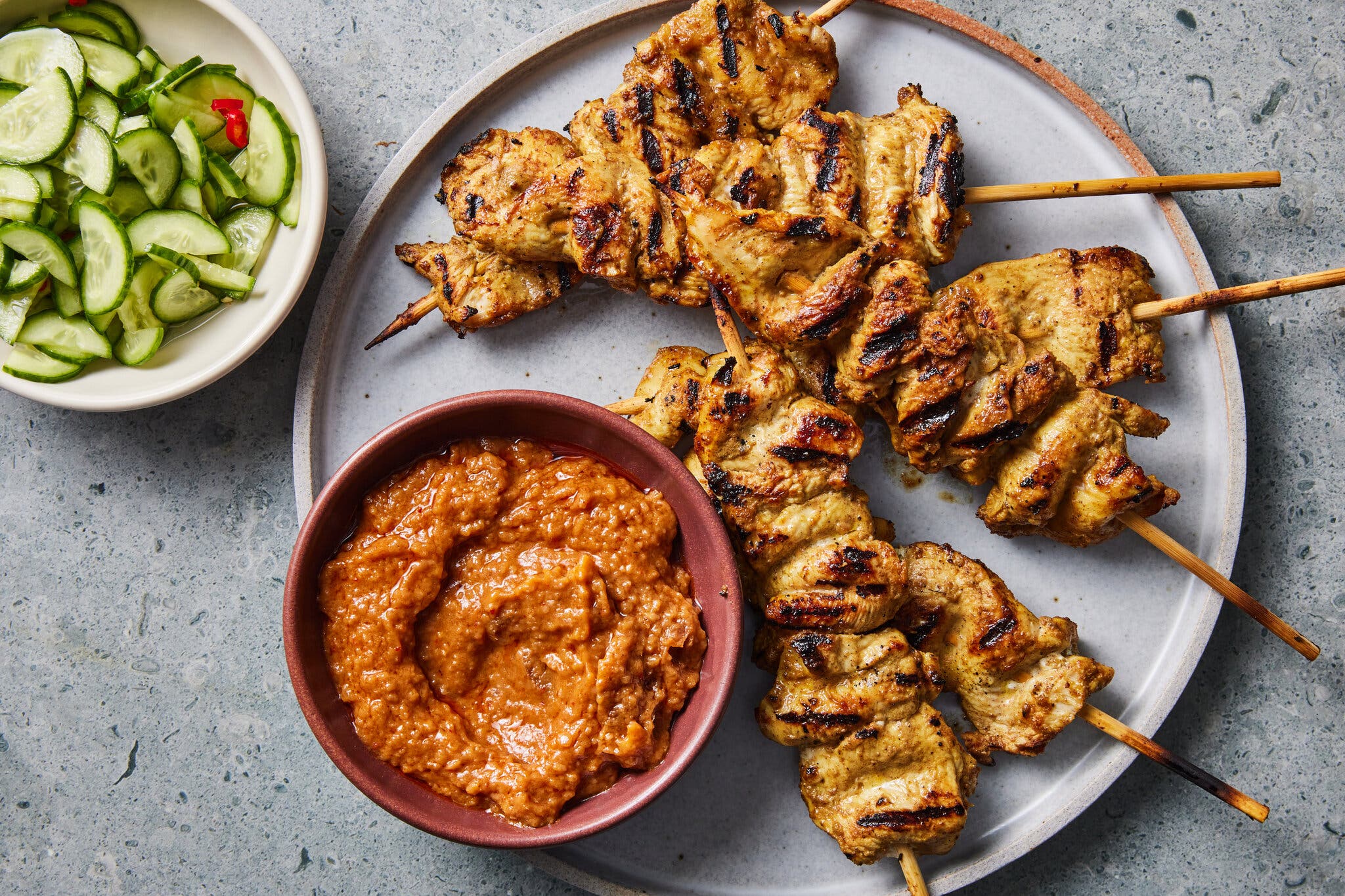 Chicken skewers lay on a plate next to a deep red-orange sauce next to a small bowl of cucumbers.