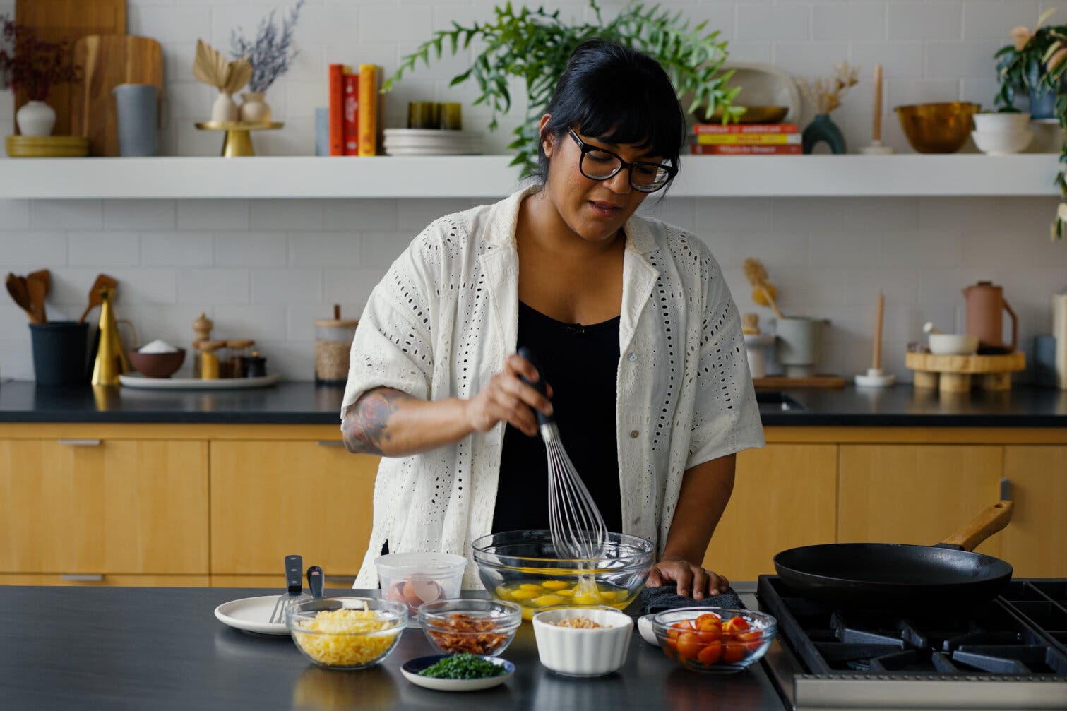 A picture of a woman with bangs and glasses standing at a kitchen counter. She is holding a whisk above a bowl of eggs. 