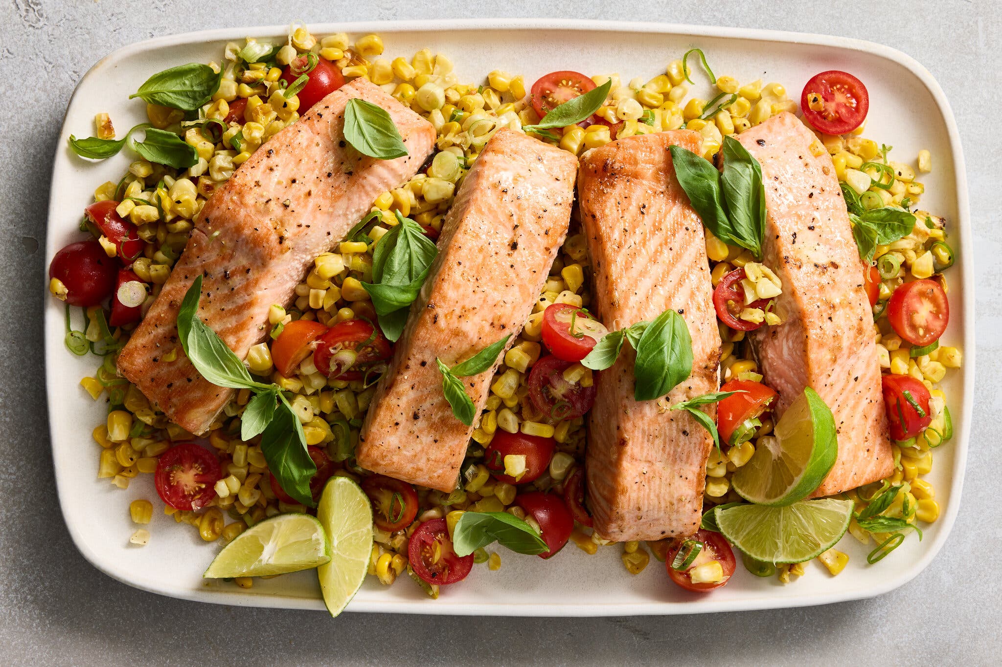 An overhead image of salmon fillets on a white tray, surrounded by corn kernels and tomatoes.