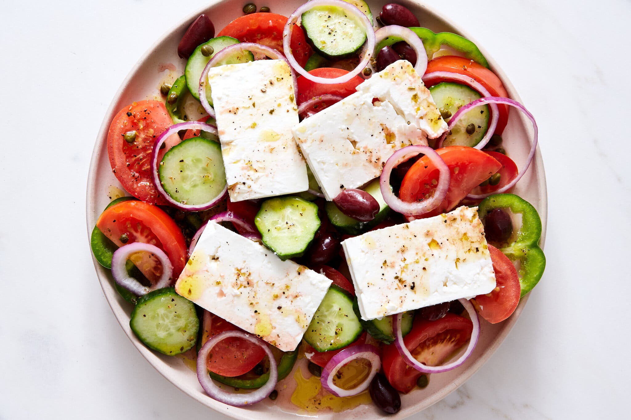 An overhead image of a platter of tomatoes and cucumbers topped with slabs of feta and rings of red onion.