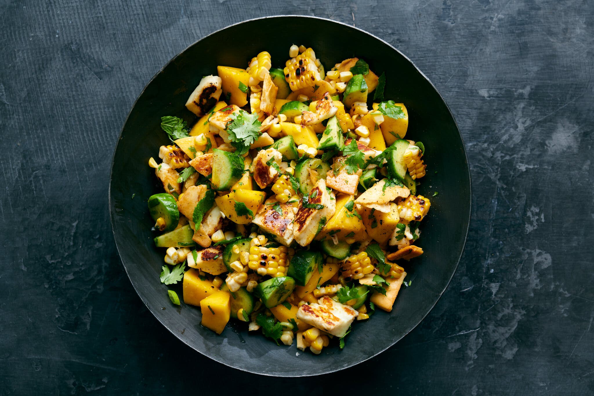 An overhead image of corn and mango tossed with cucumbers and halloumi in a black bowl.