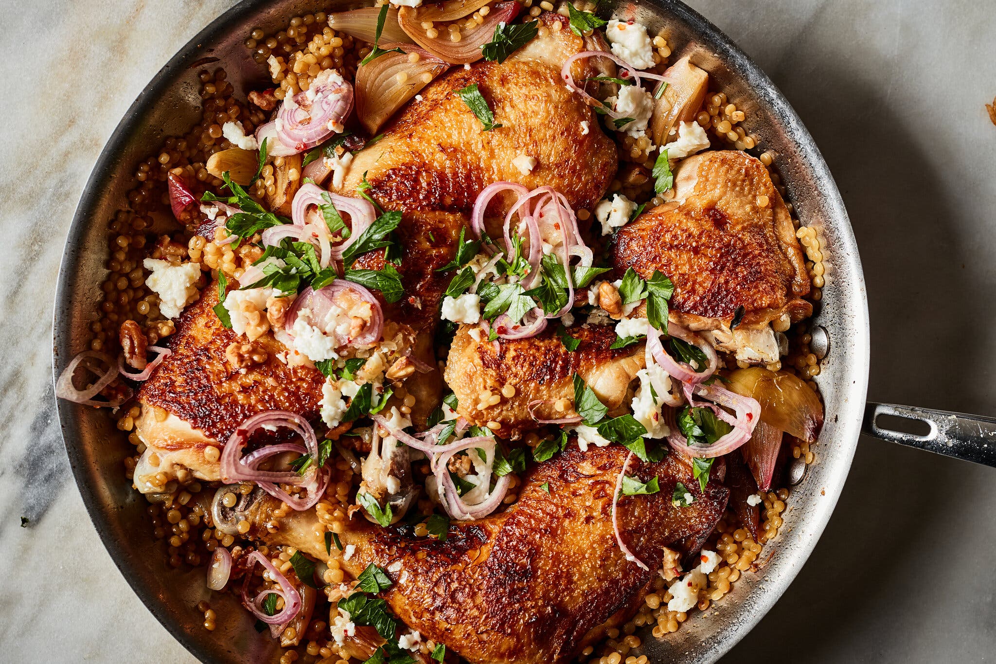 A skillet filled with burnished chicken parts is topped with shallot rings, cheese and herbs.