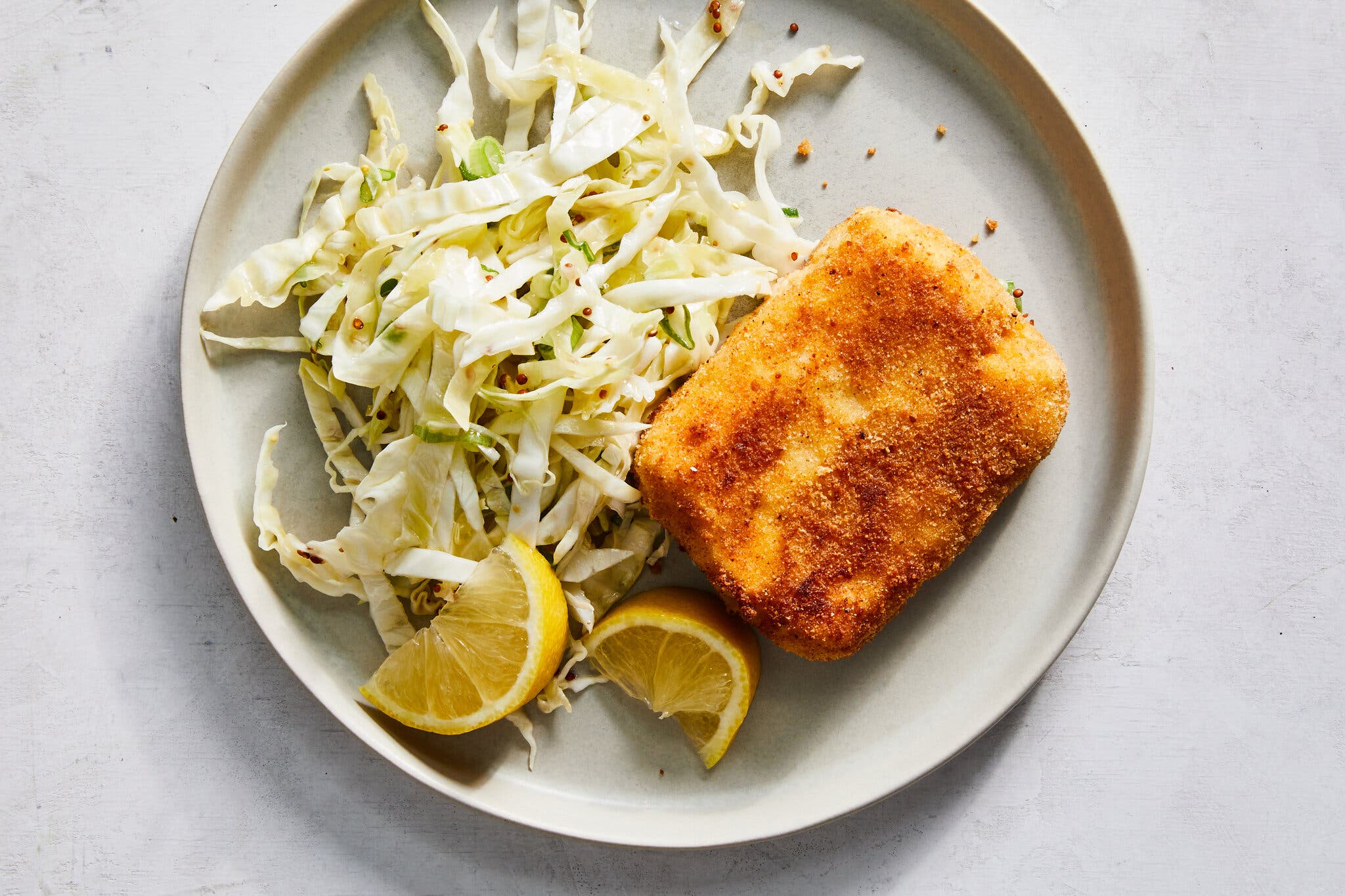 An overhead image of a breaded block of halloumi and a small cabbage slaw on a plate next to spent lemon wedges.
