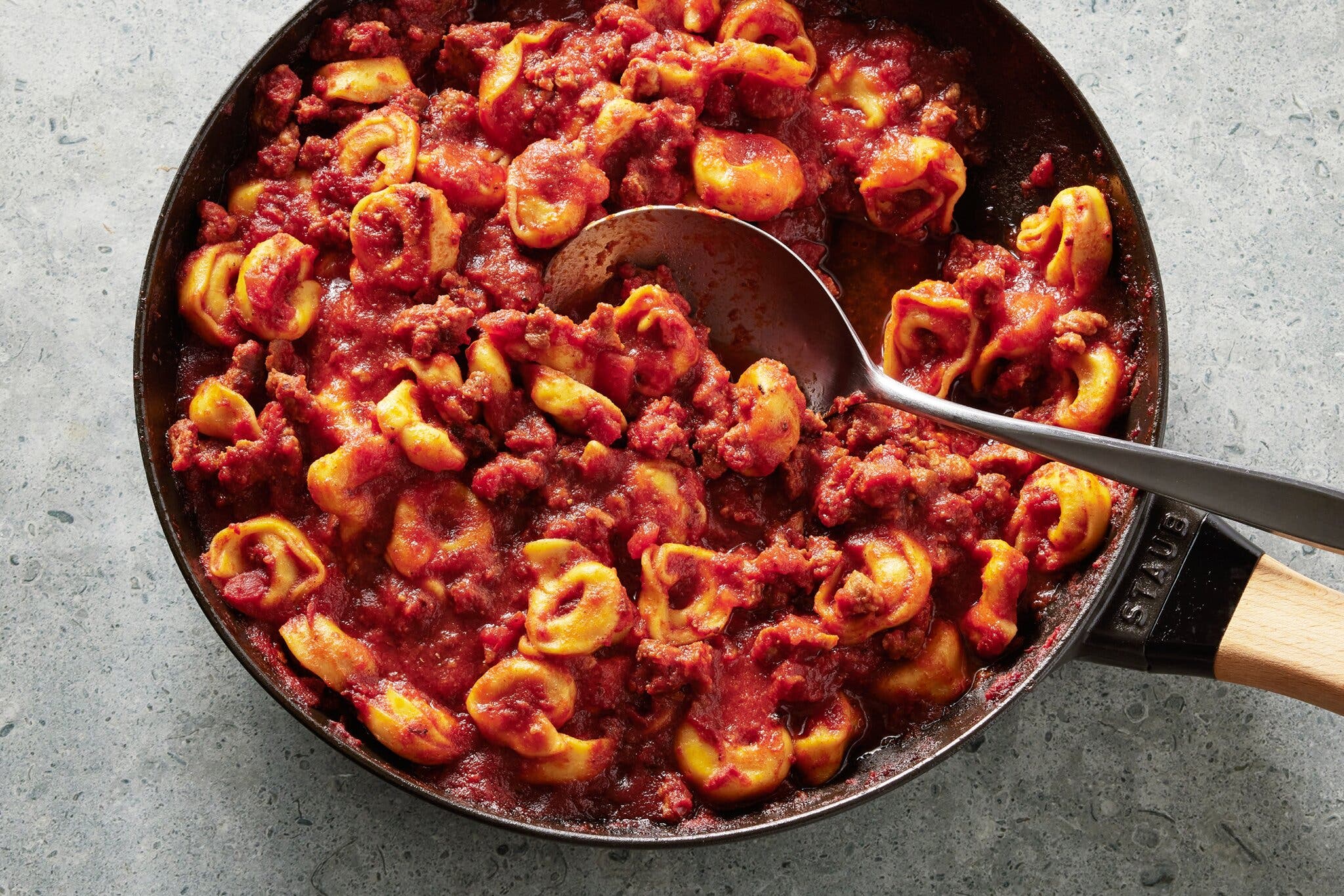 A metal spoon is scooping into a skillet of tortellini that’s been tossed with a red meat sauce.