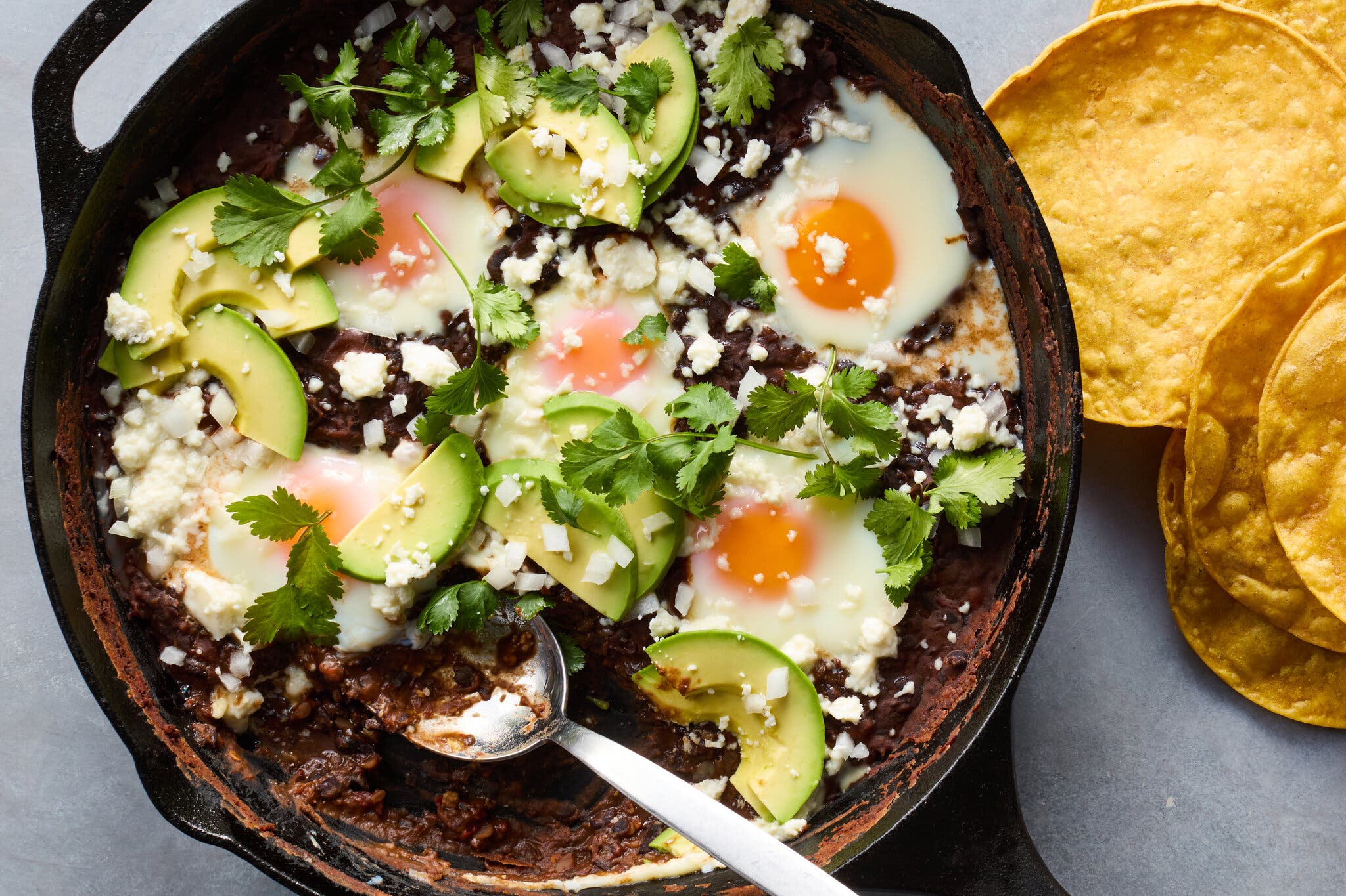 An overhead image of a cast-iron skillet filled with beans topped with eggs, sliced avocado and herbs.