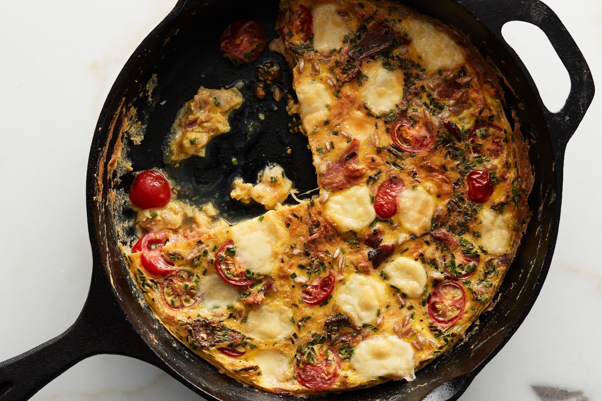 An overhead shot of a cast-iron skillet with a frittata with pockets of melted cheese and blistered cherry tomatoes. A wedge is cut out of the frittata.