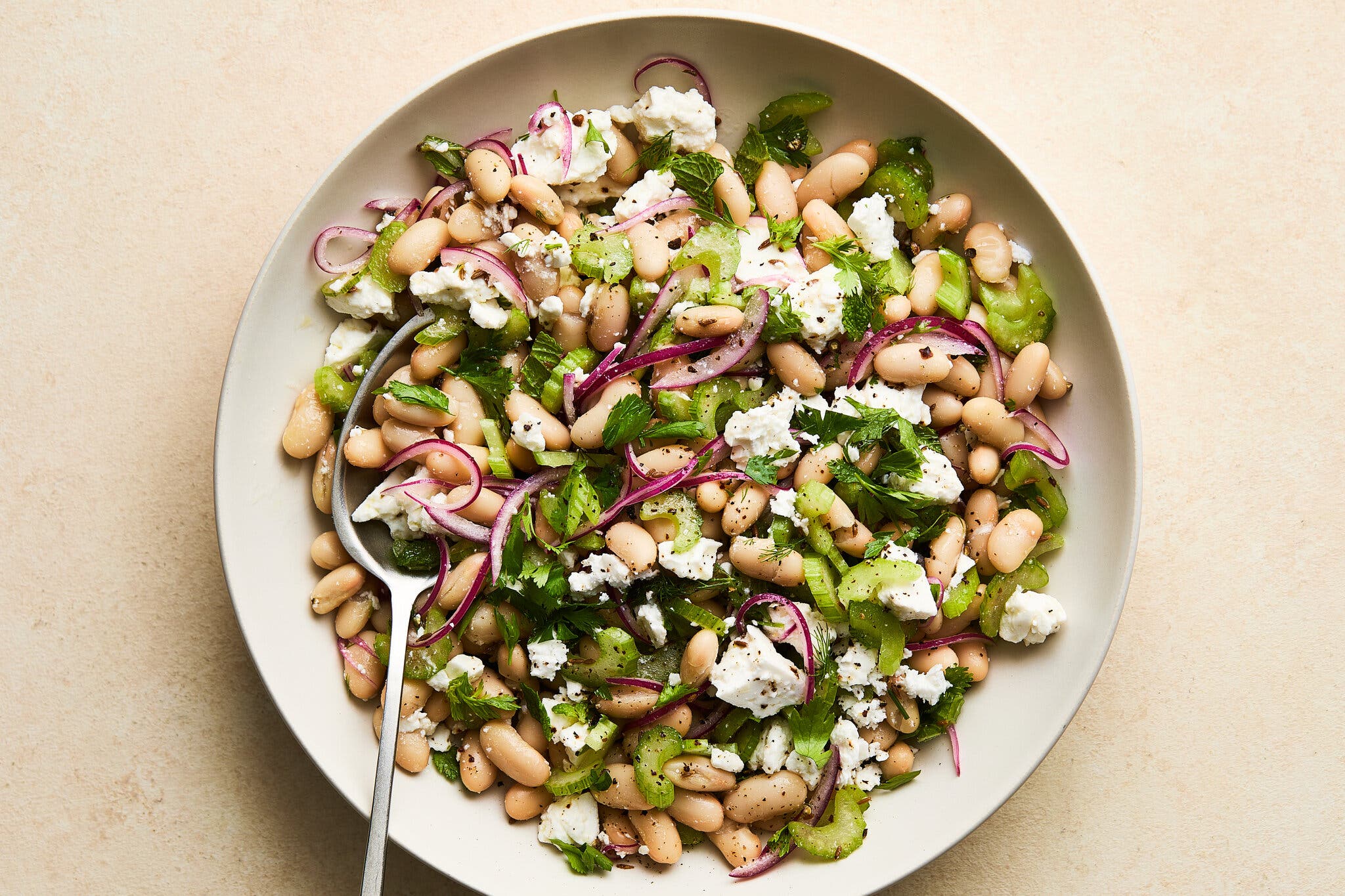 An overhead image of a white plate filled with white bean salad with sliced red onions, feta cheese, celery and parsley.