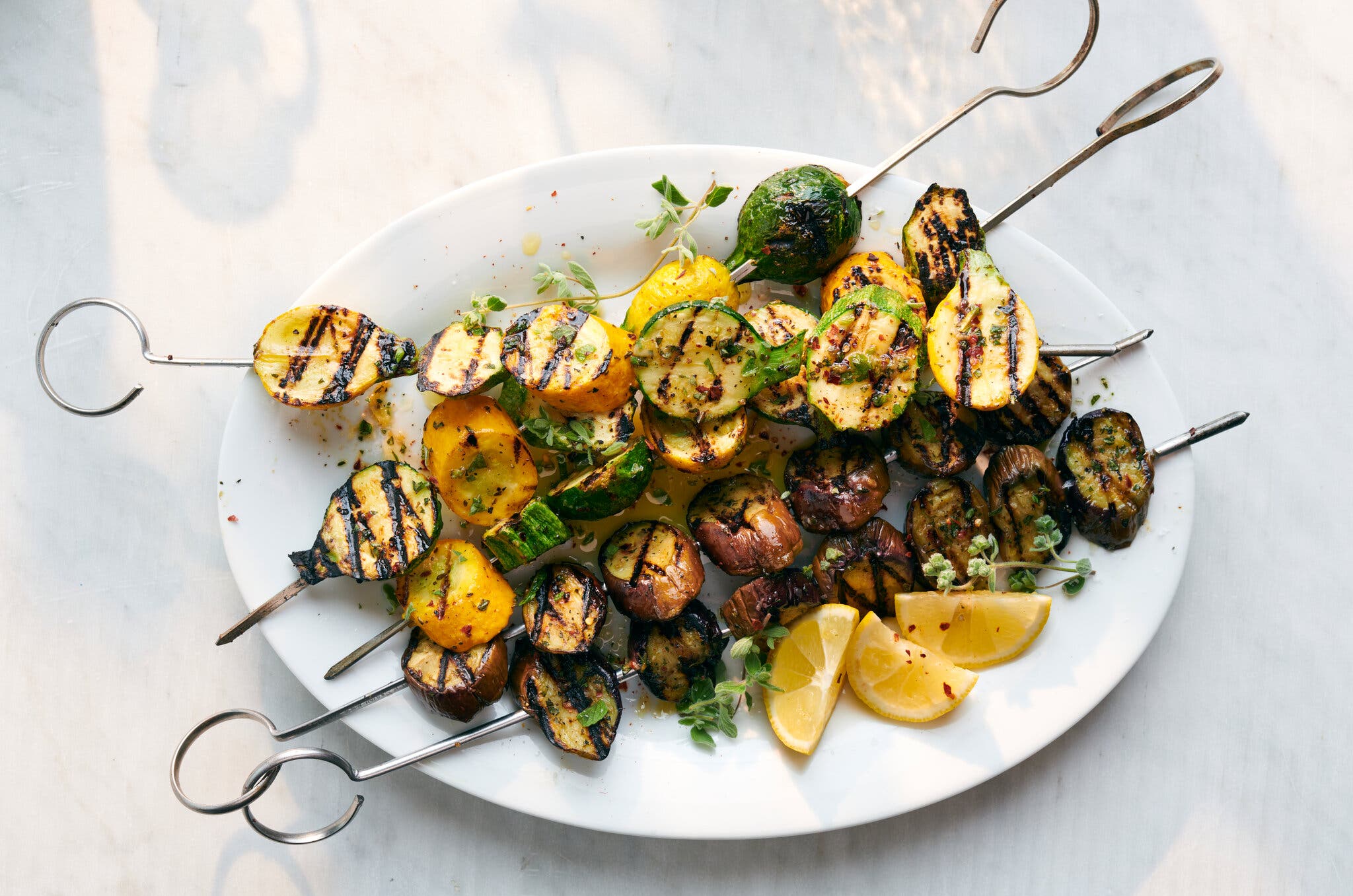 An overhead shot of a white plate full of metal skewers of grilled vegetables, like zucchini and eggplant, as well as wedges of lemon for serving.