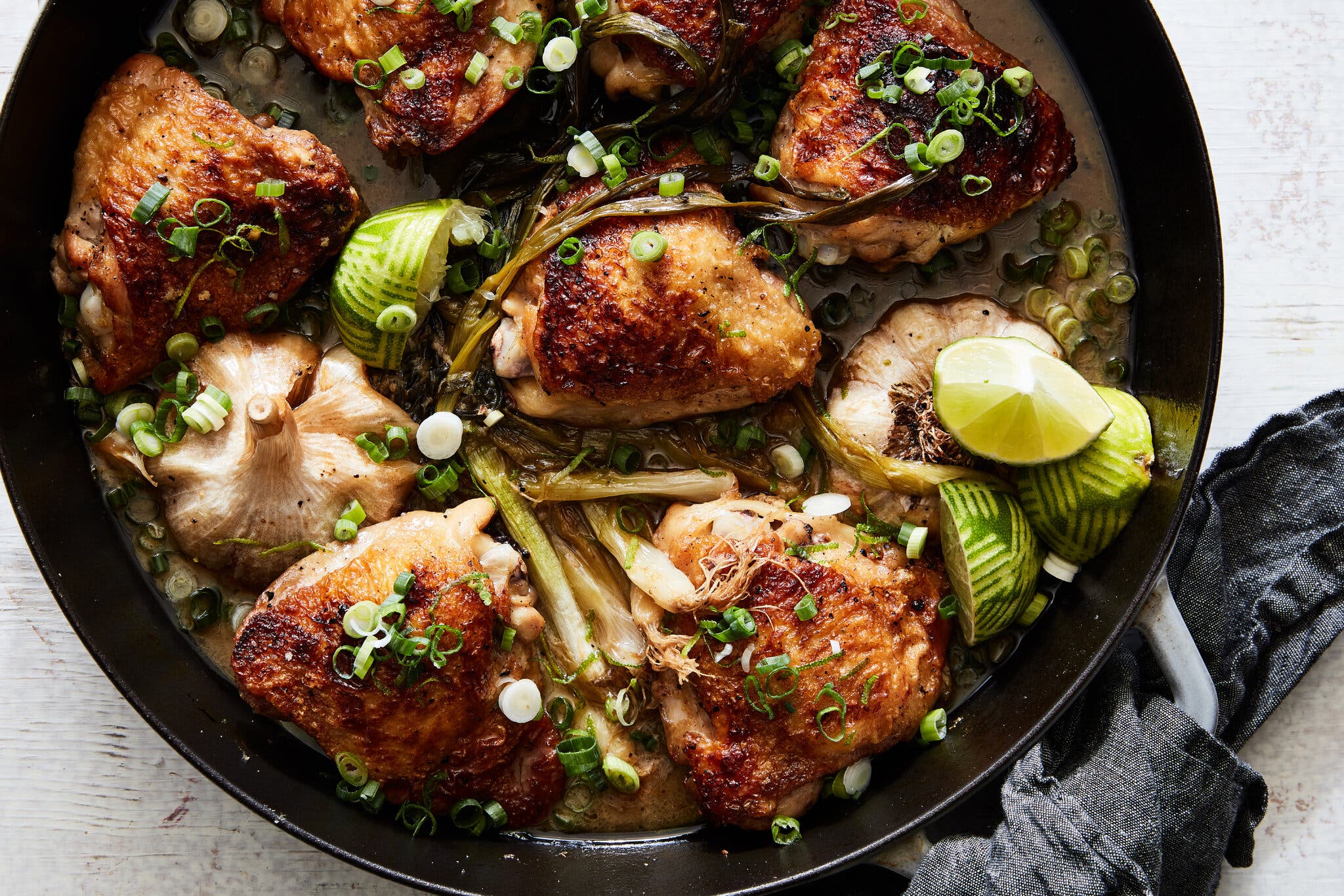 An overhead image of a cast-iron skillet with chicken thighs, a whole bulb of garlic, scallions and lime.