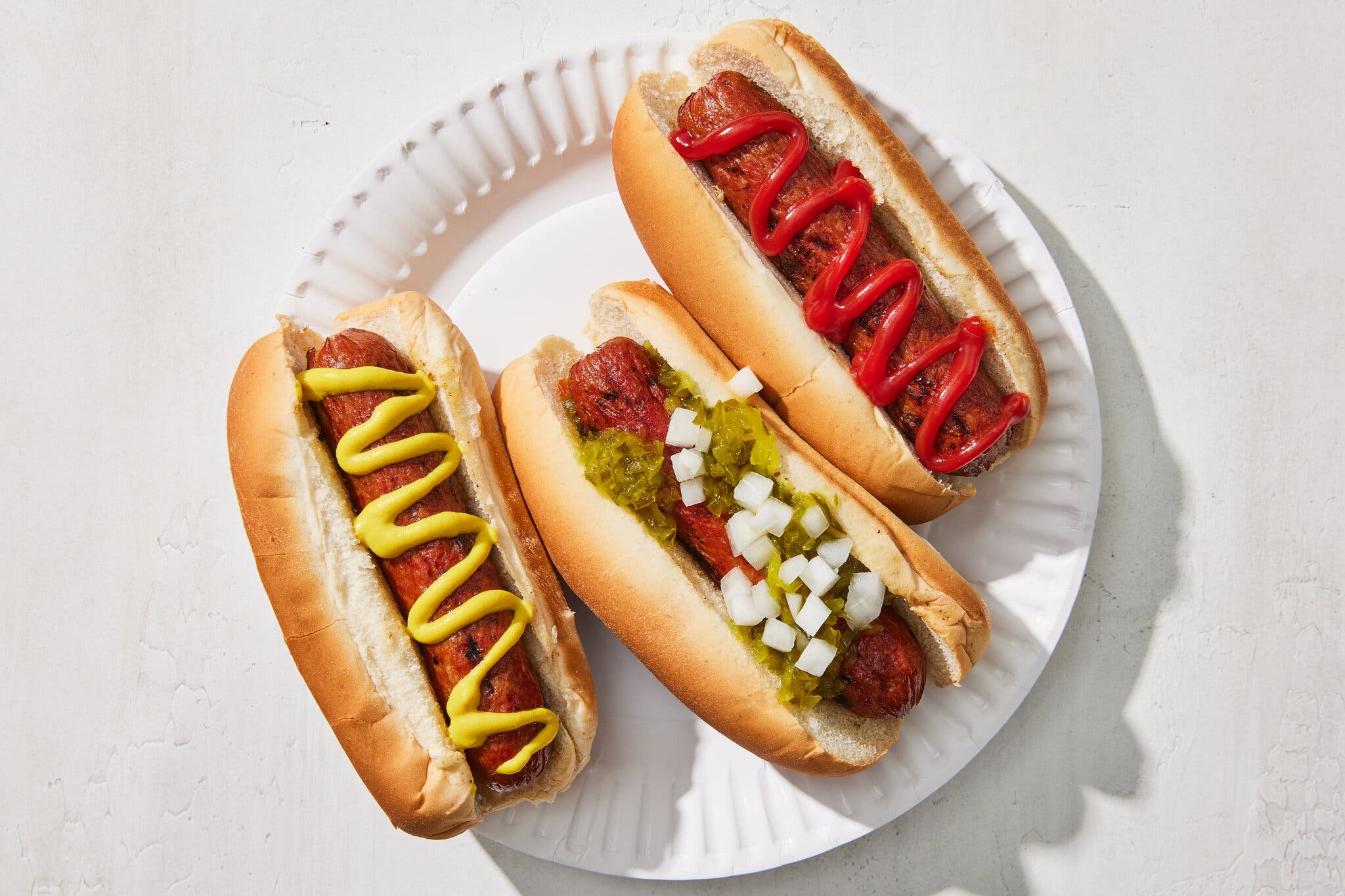 An overhead shot of three hot dogs on a white plate with crinkled edges. One is drizzled with ketchup, one with mustard and one with relish and diced white onion.