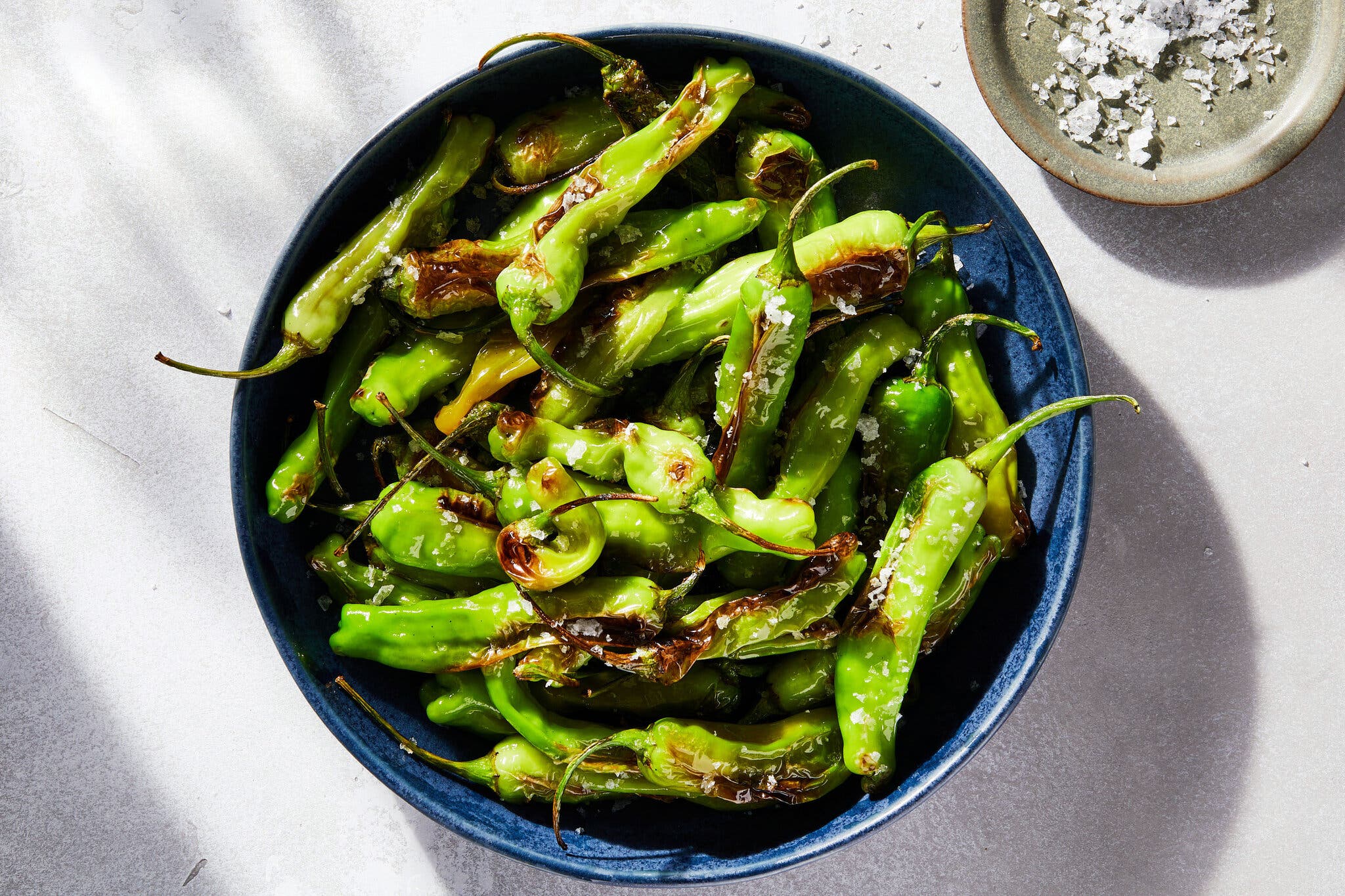 An overhead image of a blue bowl full of blistered shishito peppers. A small bowl of flaky salt is visible in the top right corner.