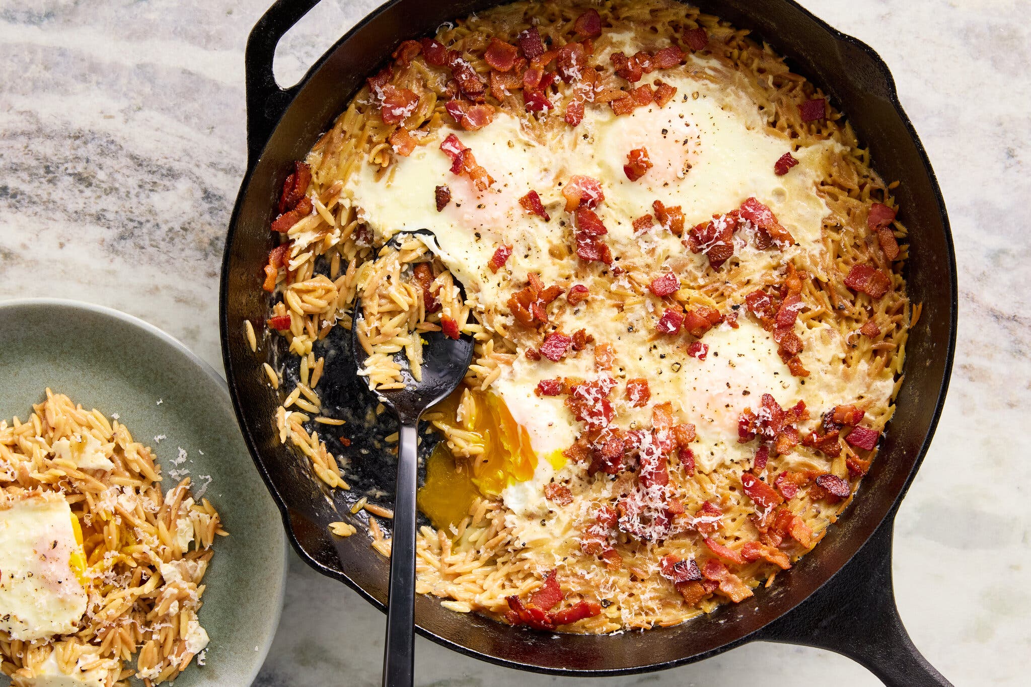 An overhead image of a cast-iron skillet full of orzo with bacon and eggs on top and a spoon inside. To the left, there is a smaller plate of orzo.