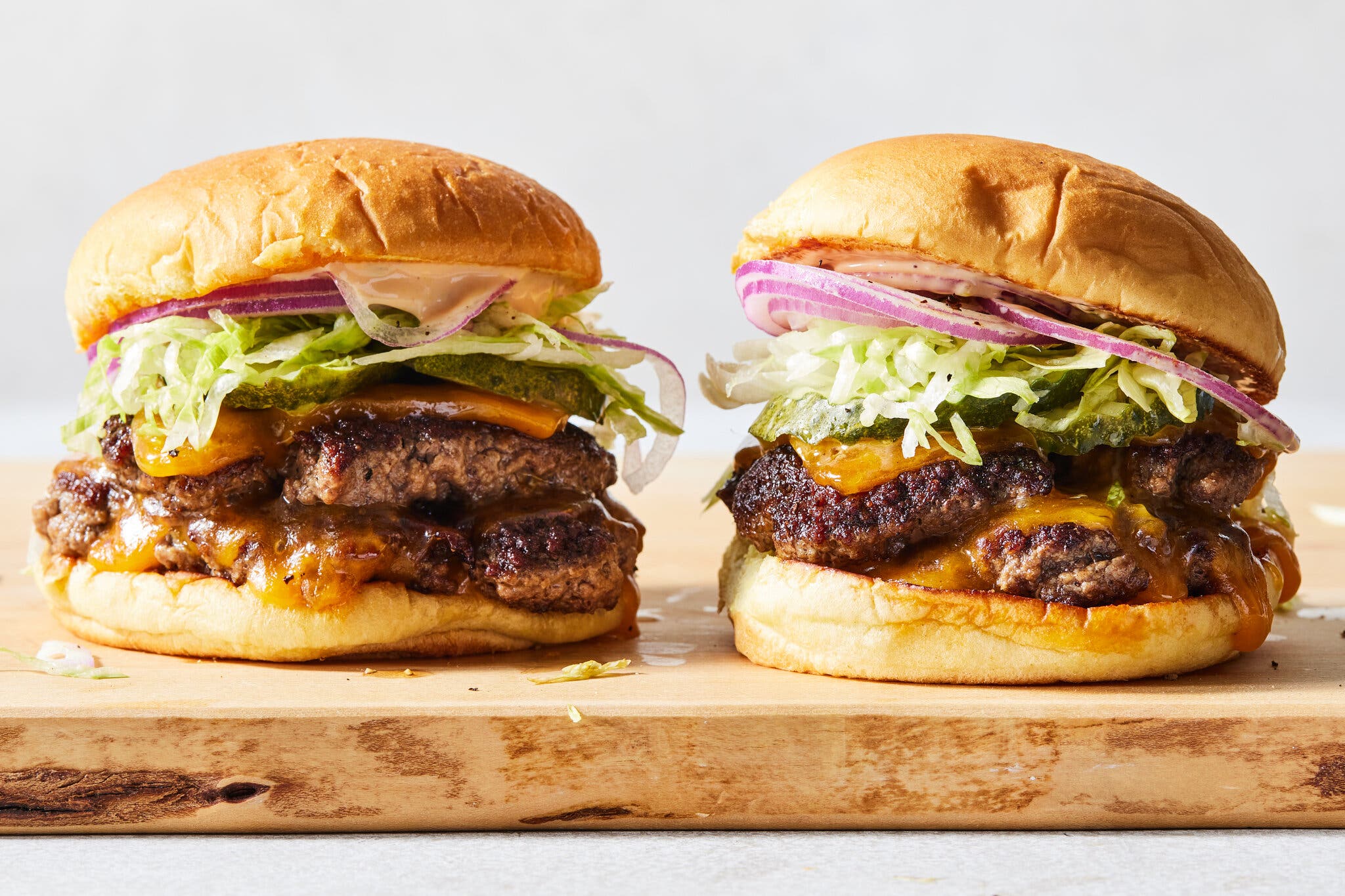 An image shot from the side of two burgers on a wooden cutting board. The burgers have two patties each, melted Cheddar cheese, shredded lettuce, red onion and mayonnaise.