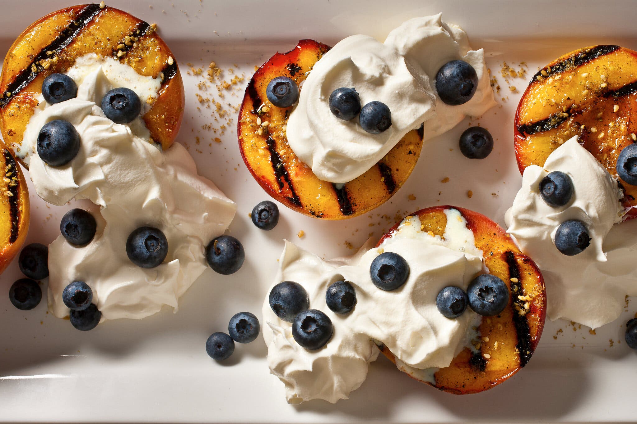 Five grilled peaches with blueberries and whipped cream are topped with dukkah and arranged on a rectangular white platter.