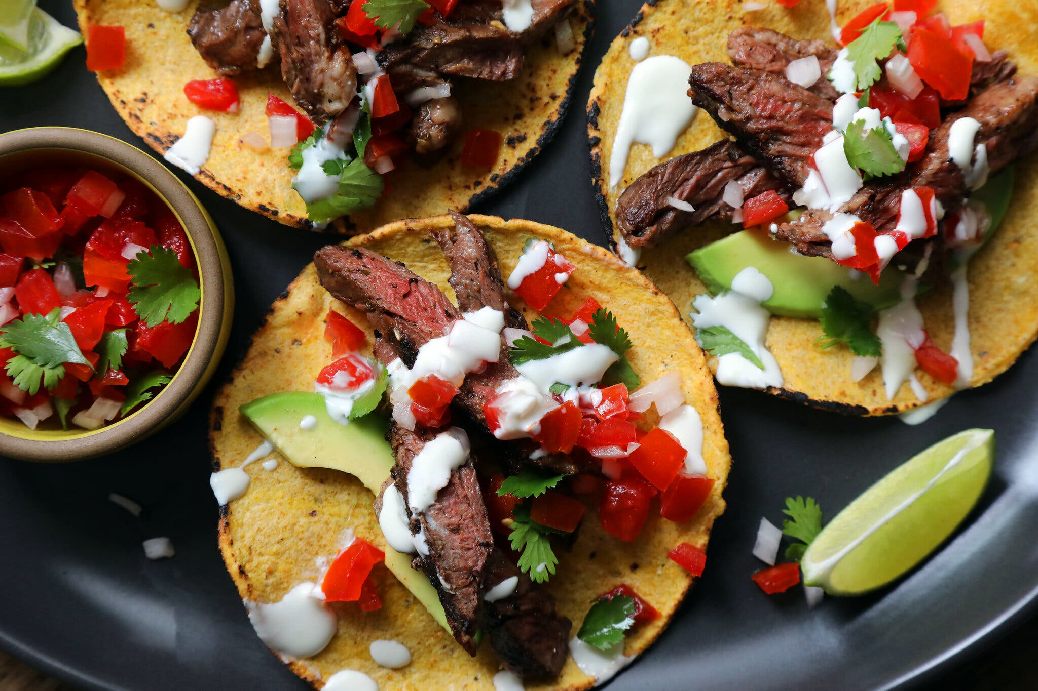 An overhead image of three corn tacos on a black plate. The tacos have carne asada, sliced avocado, diced tomato, diced onion and a white sauce. A wedge of lime and small bowl of pico de gallo on the side.