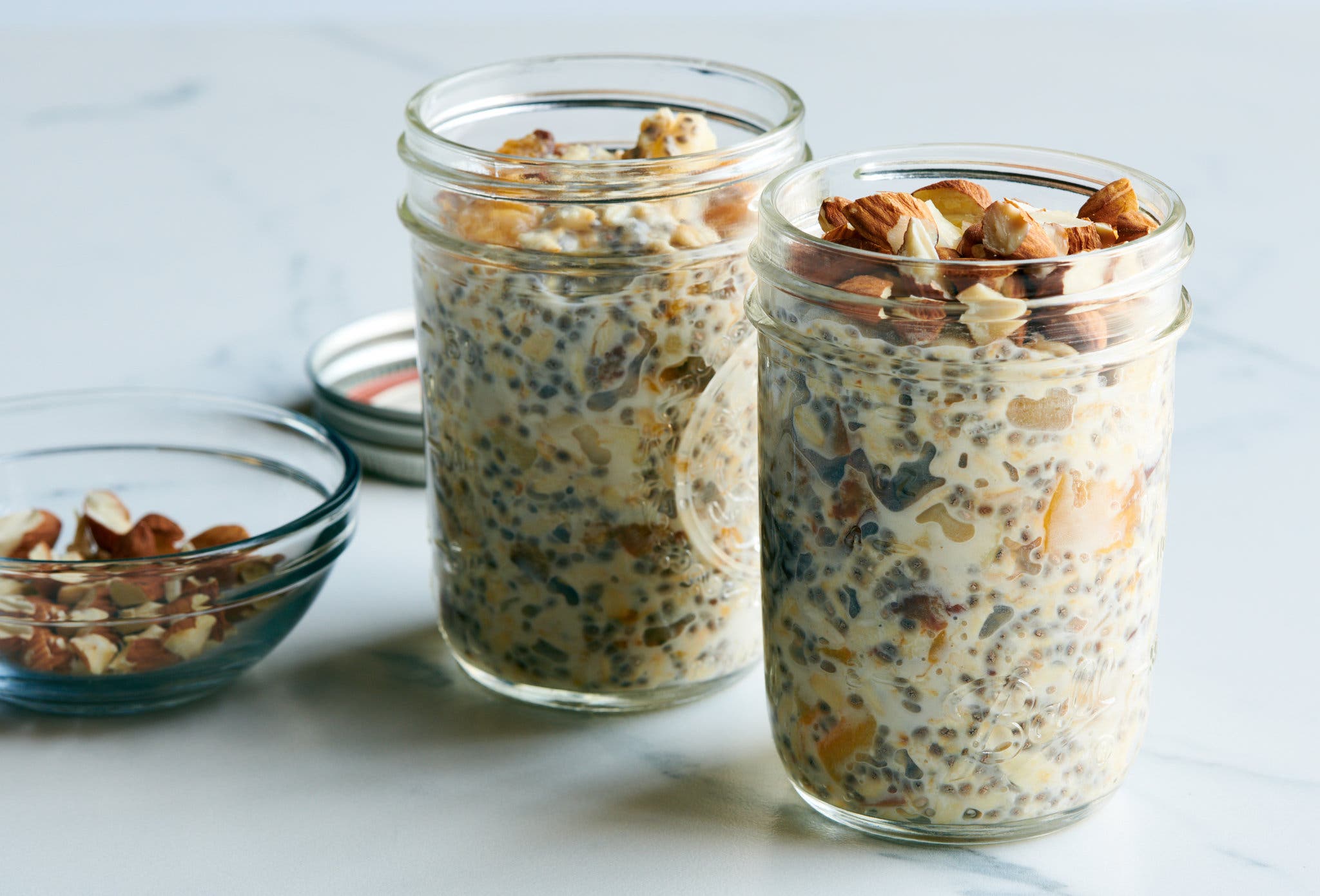 Two jars filled with overnight oats, including one topped with raw almonds, sit on a white countertop. To the left a bowl of more raw almonds is visible while a lid can be seen behind one of the jars.
