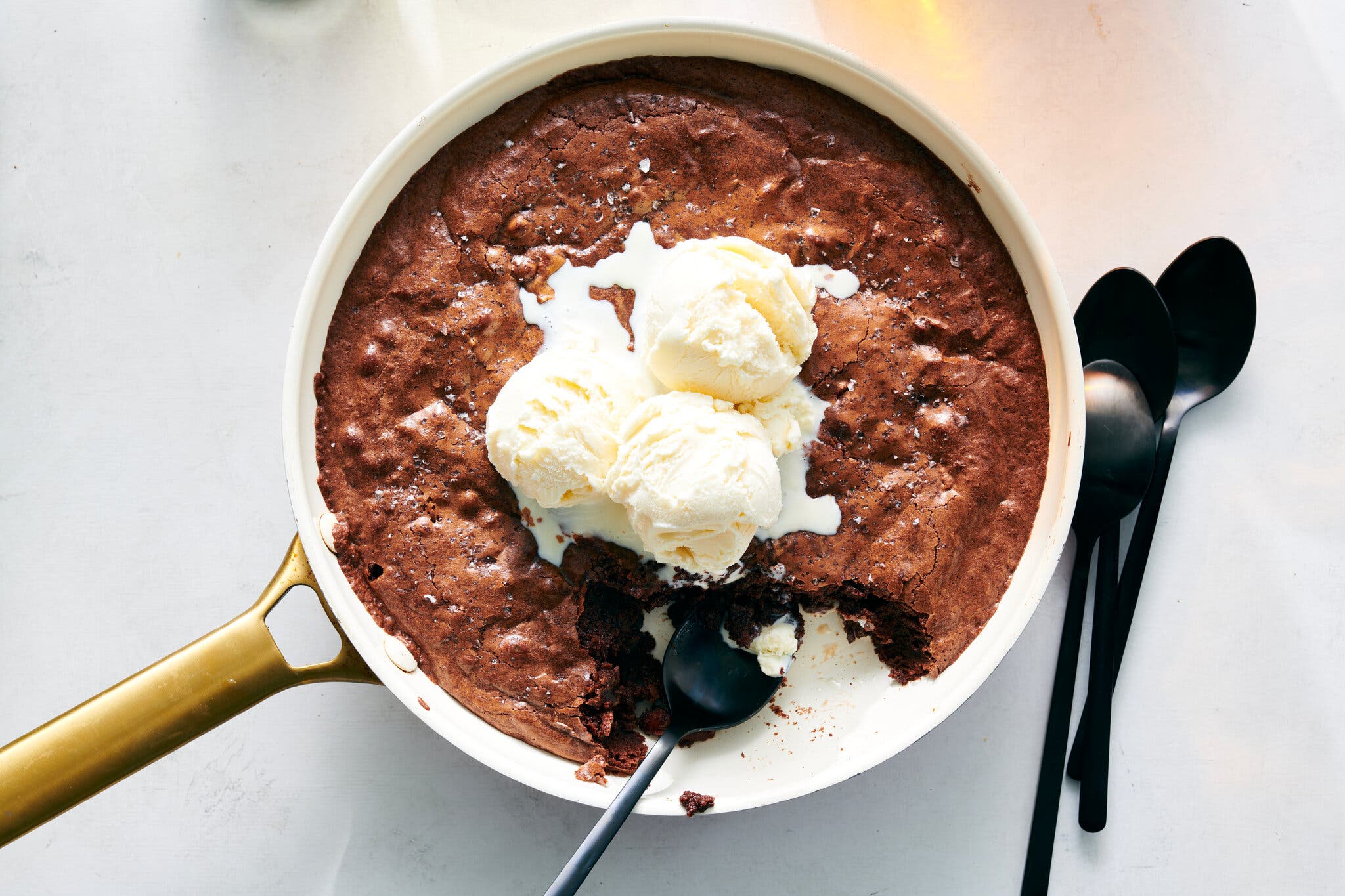 An overhead image of a skillet full of brownie, with three scoops of vanilla ice cream on top. A black spoon is in the skillet, and three spoons sit to the right of the skillet.