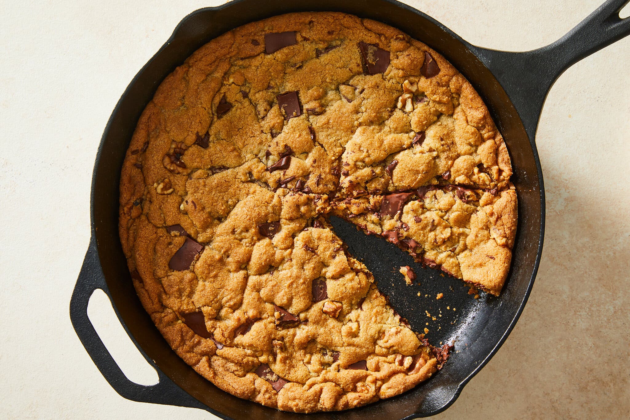 An overhead image of a cast-iron skillet with a giant chocolate chip cookie inside. A slice is cut out of the cookie.