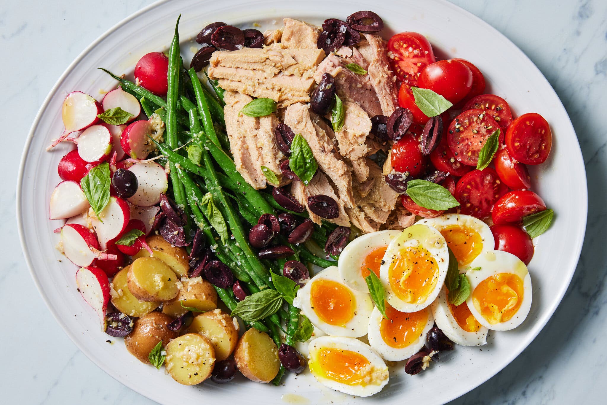 An overhead image of a platter topped with tuna, tomatoes, halved hard-boiled eggs, olives, green beans, potatoes and radishes.