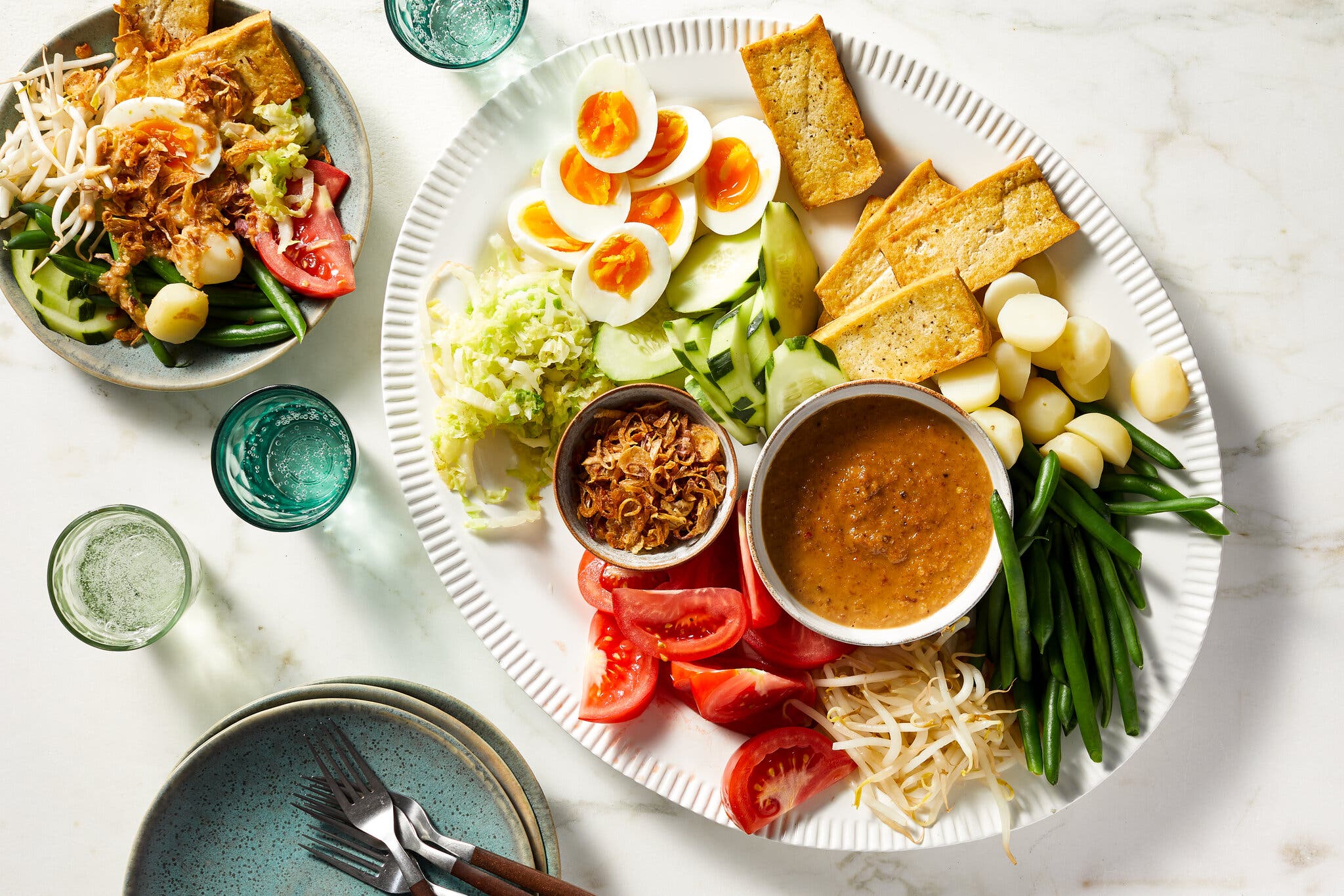 Sabs of tofu, potatoes, green beans, bean sprouts, tomatoes and halved eggs surround fried shallots and a dark brown sauce on a white platter. A single serving sits to the side.