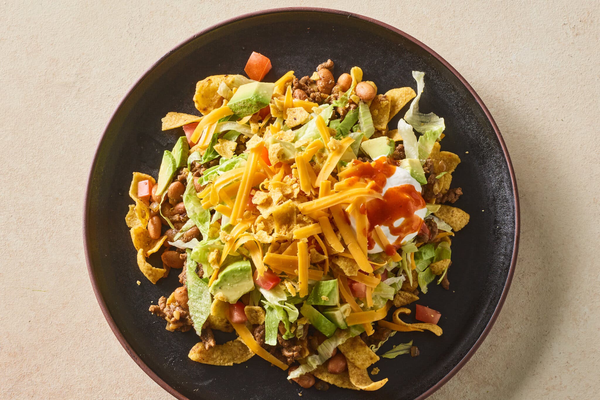 An overhead image of a black bowl topped with corn chips, lettuce, shredded cheese, sour cream and hot sauce.