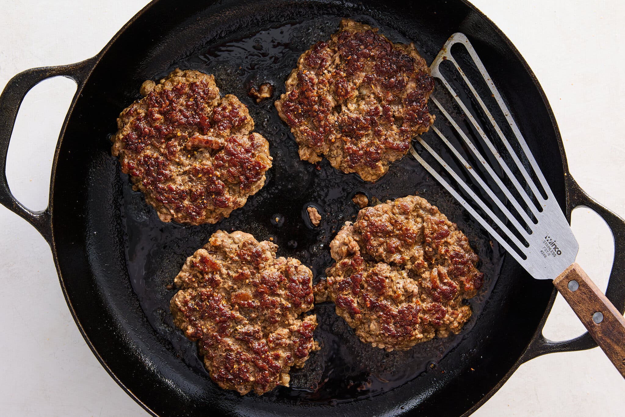 An overhead image of a cast-iron skillet filled with four browned beef patties. A spatula sits to the side.