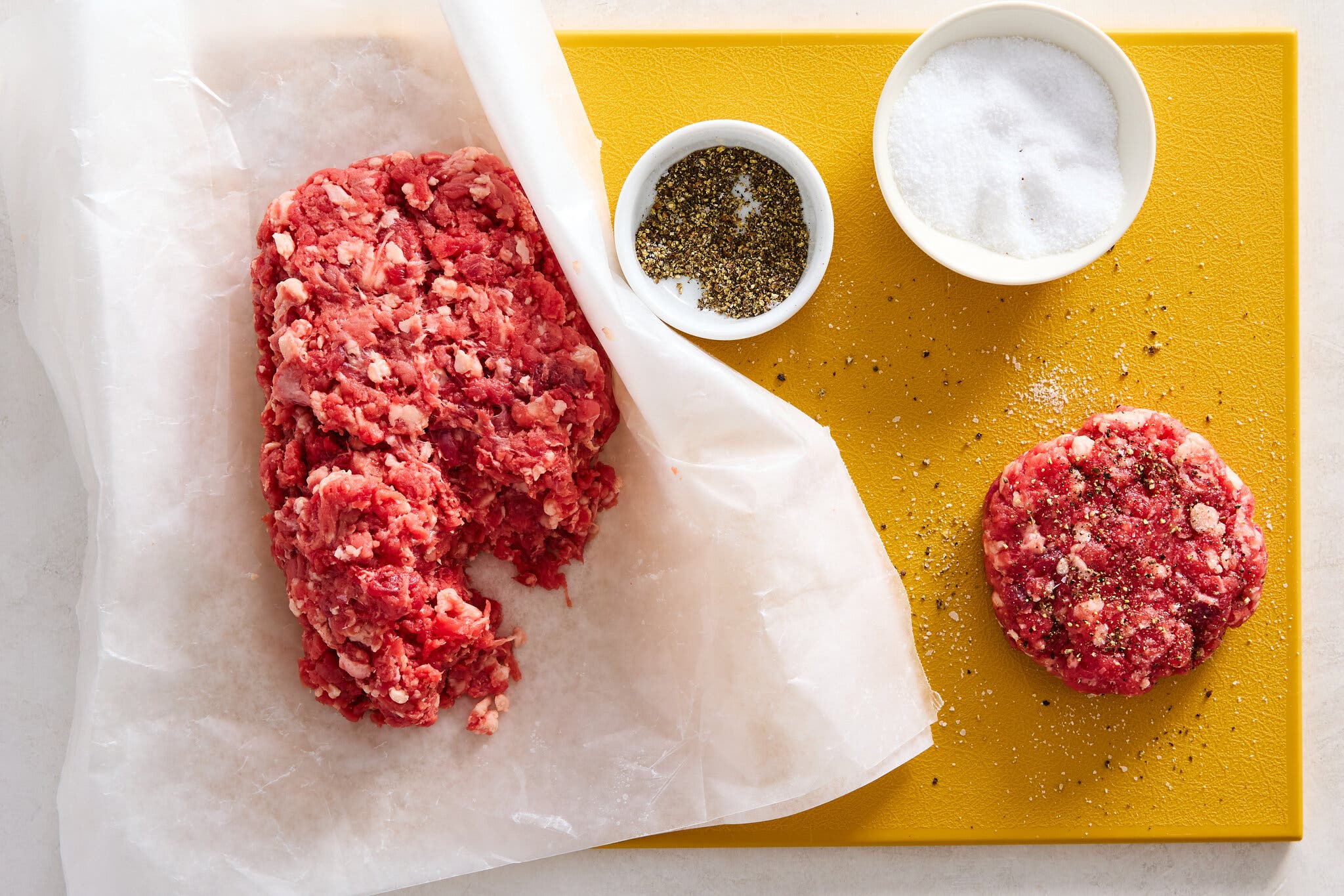 An overhead image of a slab of ground beef on a cutting board next to a single patty and bowls filled with salt and pepper.