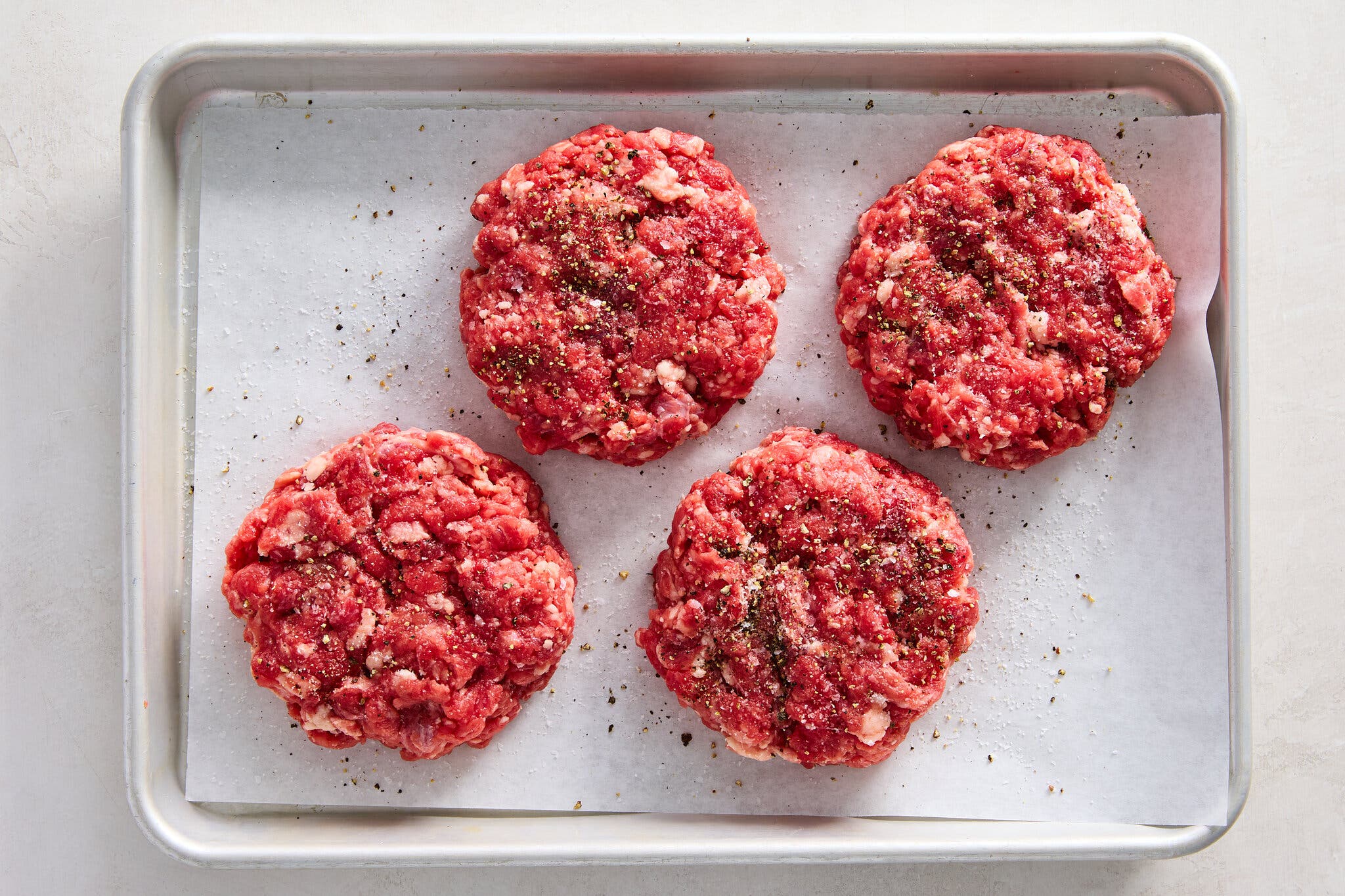 An overhead image of a small rimmed sheet pan topped with four uncooked beef patties seasoned on the outside.