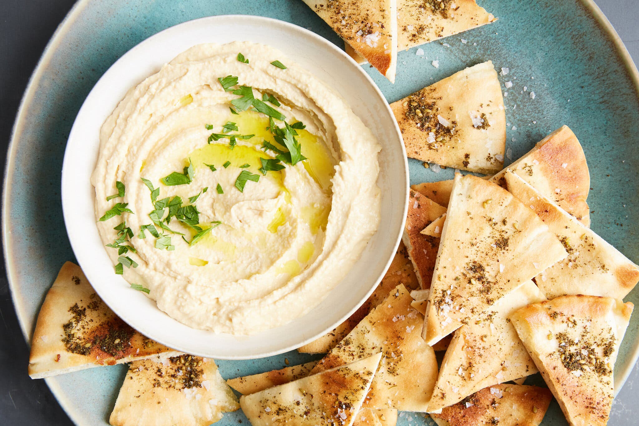 A white bowl with 5-minute hummus, topped with some olive oil and parsley, sits on a blue plate with za’atar pita chips.