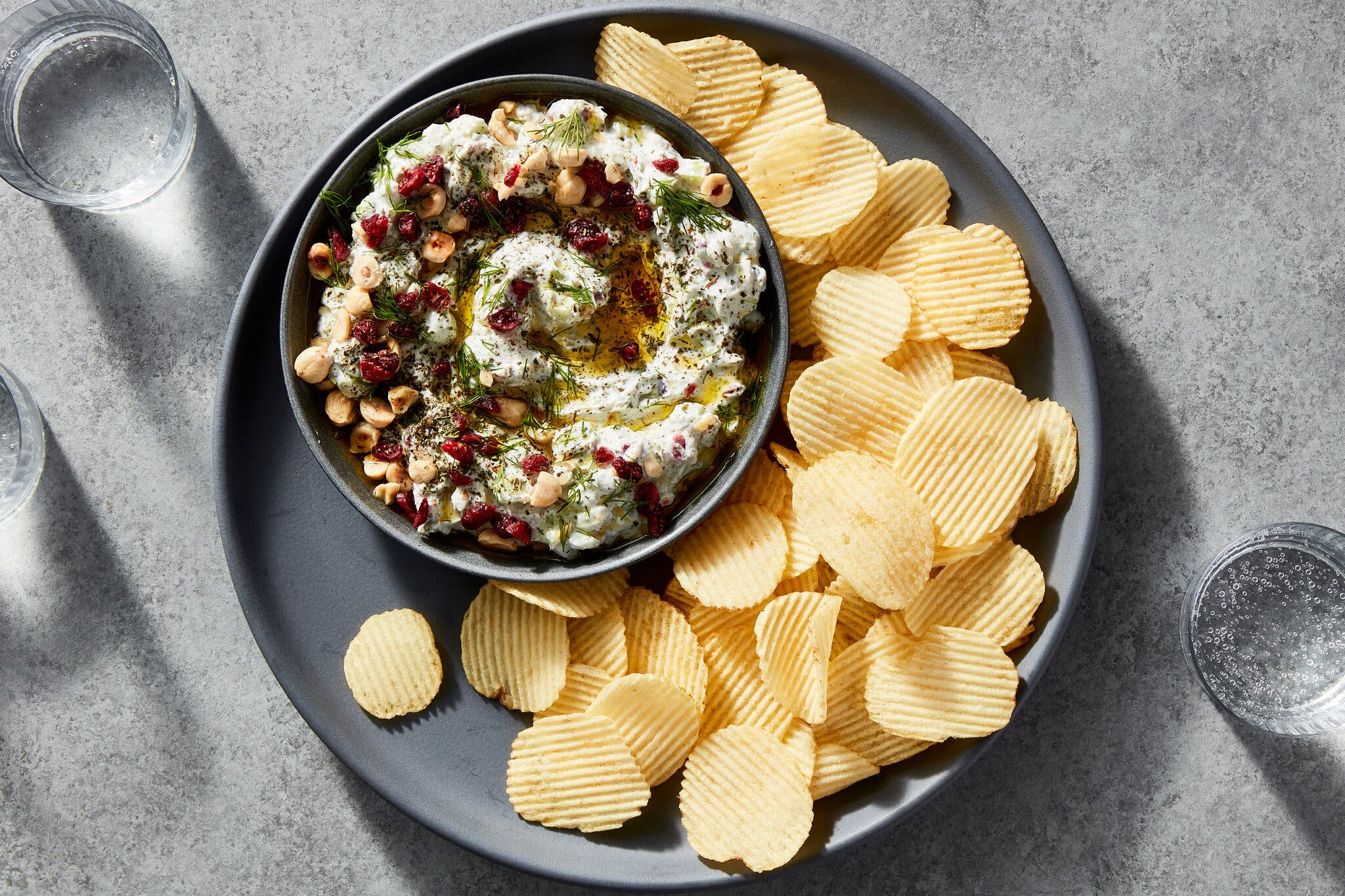 A bowl of celery yogurt dip garnished with chopped hazelnuts, dried cranberries, fresh dill and olive oil. The bowl sits on a plate with wavy potato chips. 