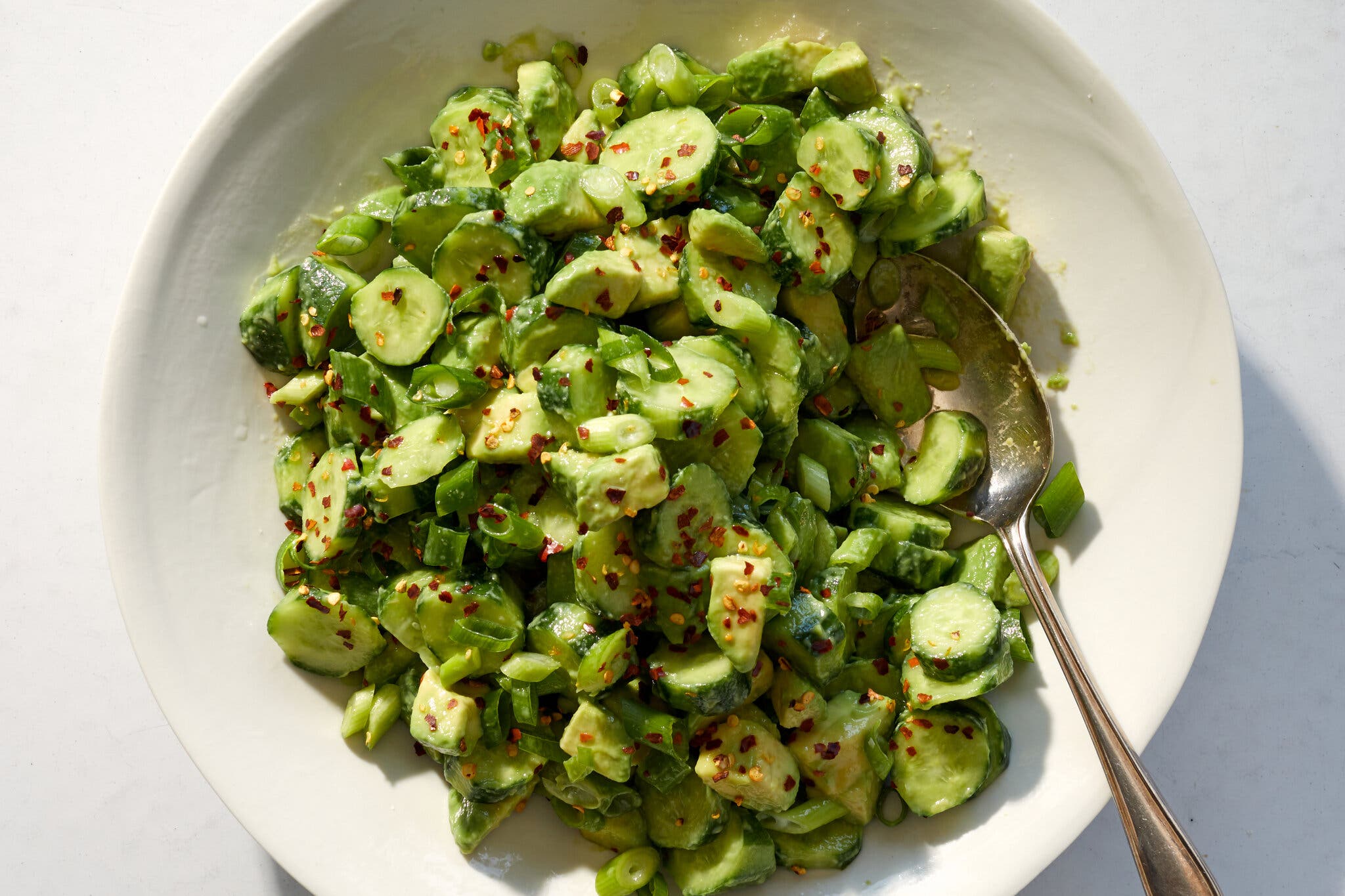 A white bowl with bite-sized pieces of cucumber and avocado, topped with chili flakes. 