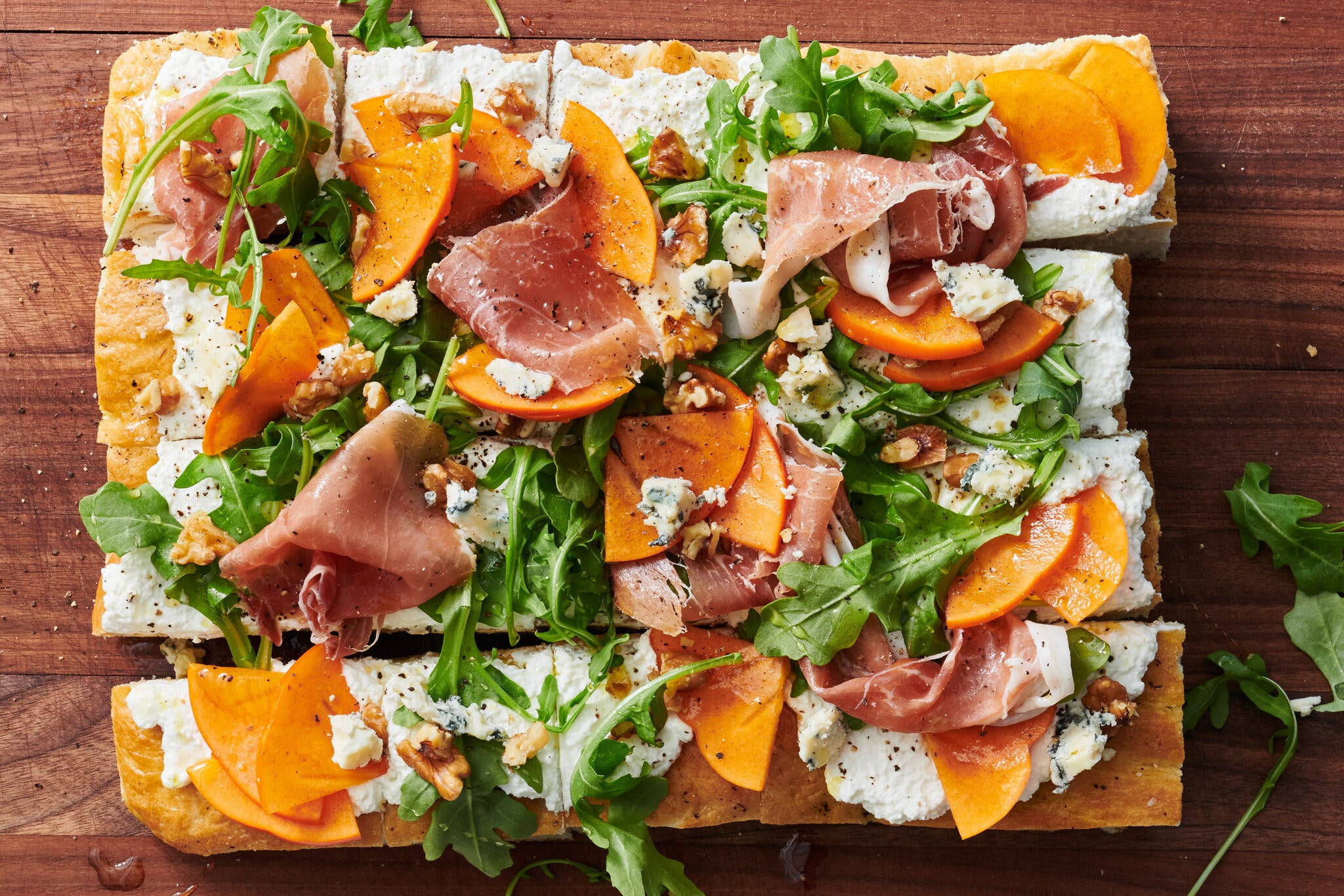 A focaccia cut into squares topped with ricotta, sliced persimmons, arugula, prosciutto and crumbled blue cheese sits on a wood cutting board. 