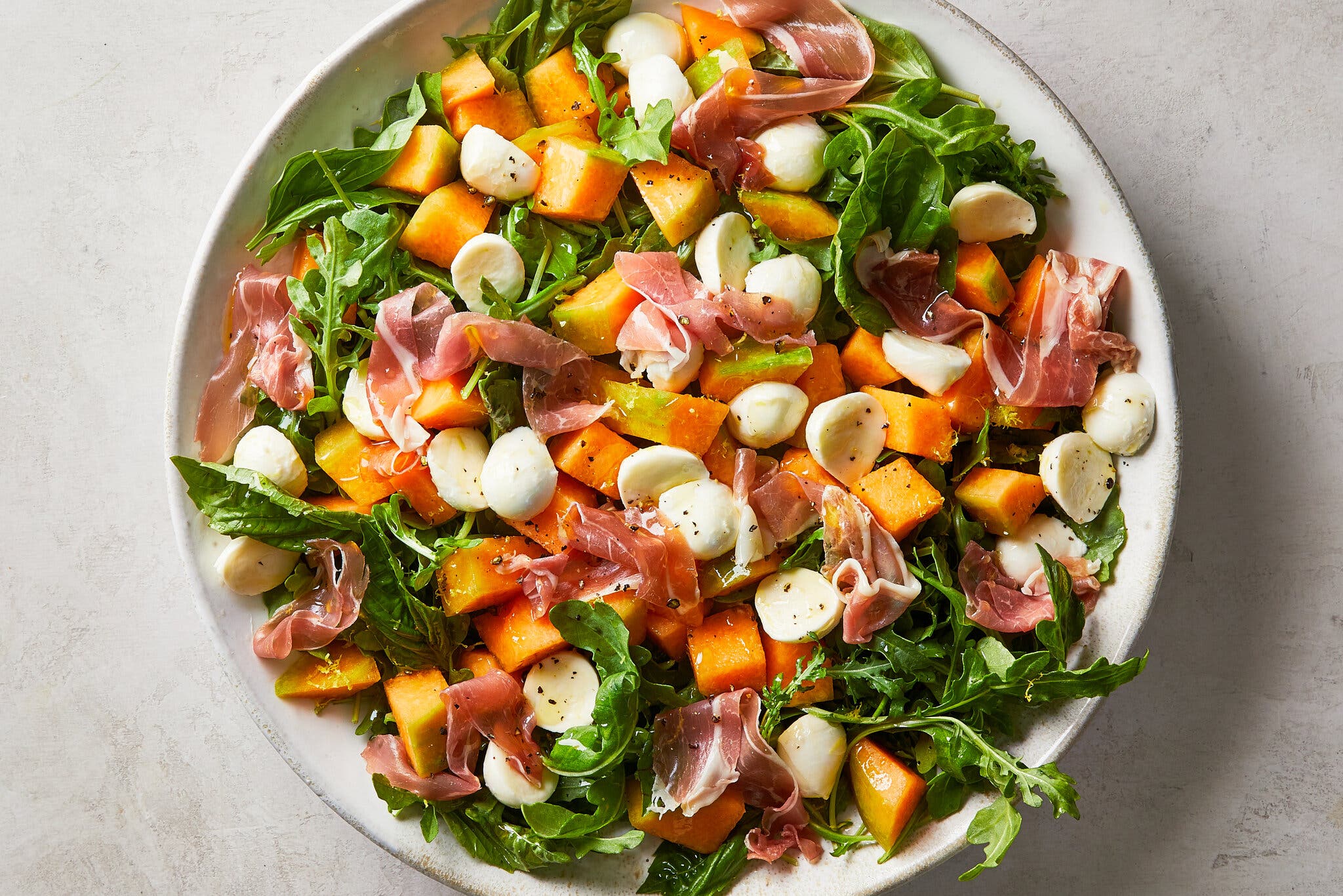 An overhead image of a big bowl of salad with arugula, cubed melon, prosciutto and small mozzarella balls cut in half. 