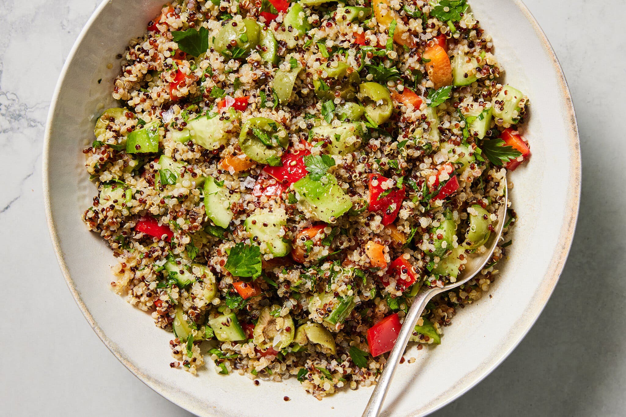 A beige bowl of quinoa salad with chopped cucumbers, bell peppers, green olives and parsley. 